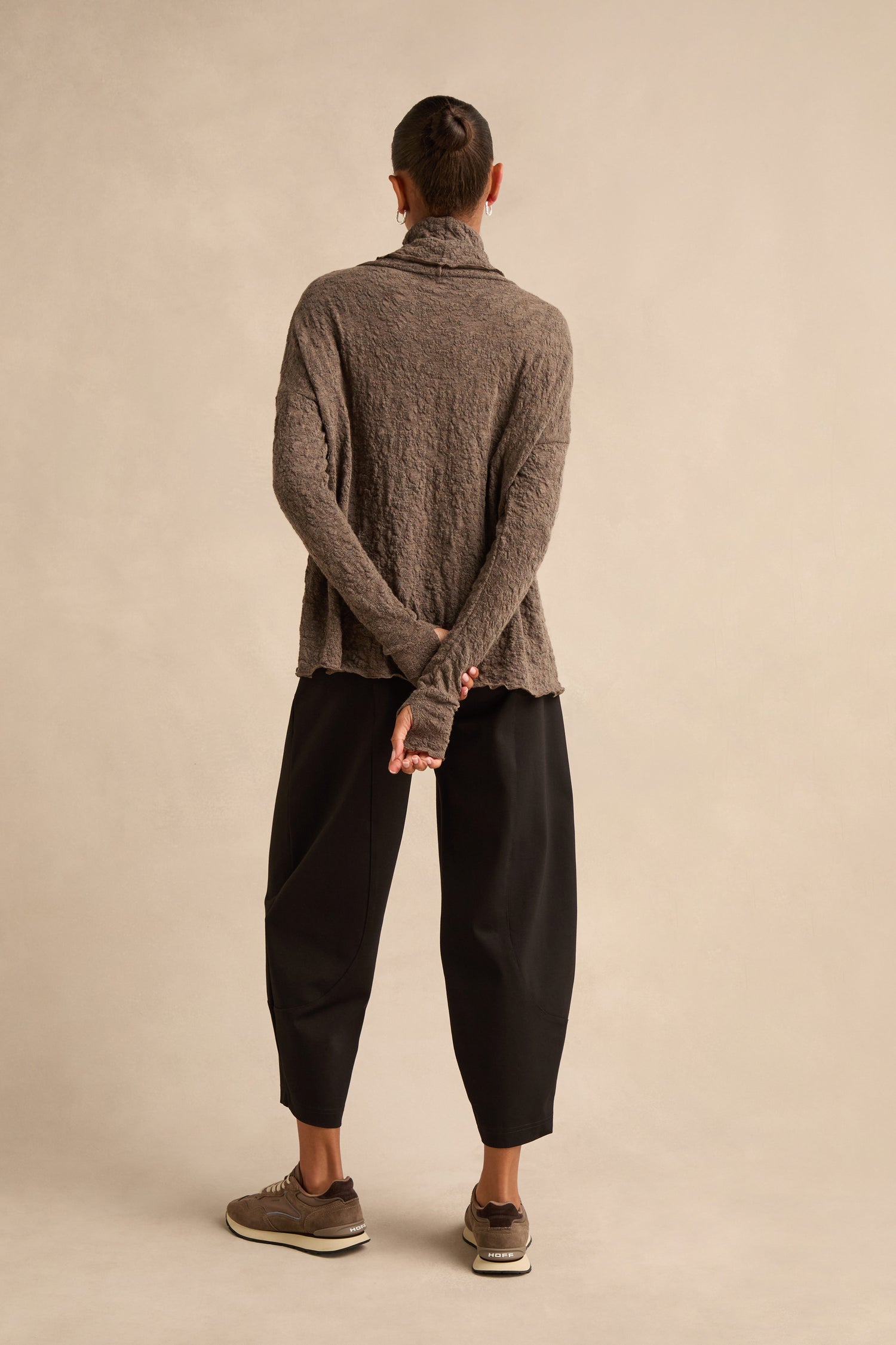 A person stands with their back to the camera, wearing the Taupe Textured Merino Roll Neck Top, paired with loose black pants and brown sneakers against a plain neutral background. Their hands are clasped behind their back.