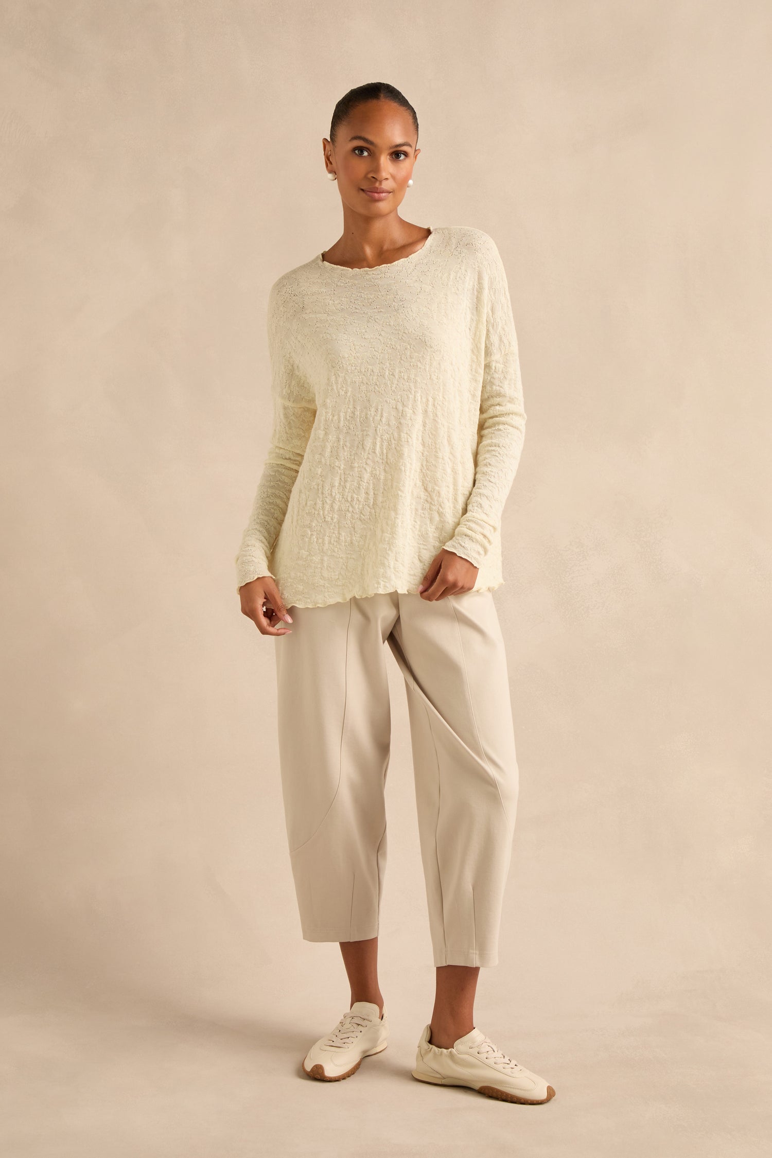 Woman standing against a beige background wearing the Ivory Textured Merino Scoop Neck Top, light beige cropped pants, and white sneakers.
