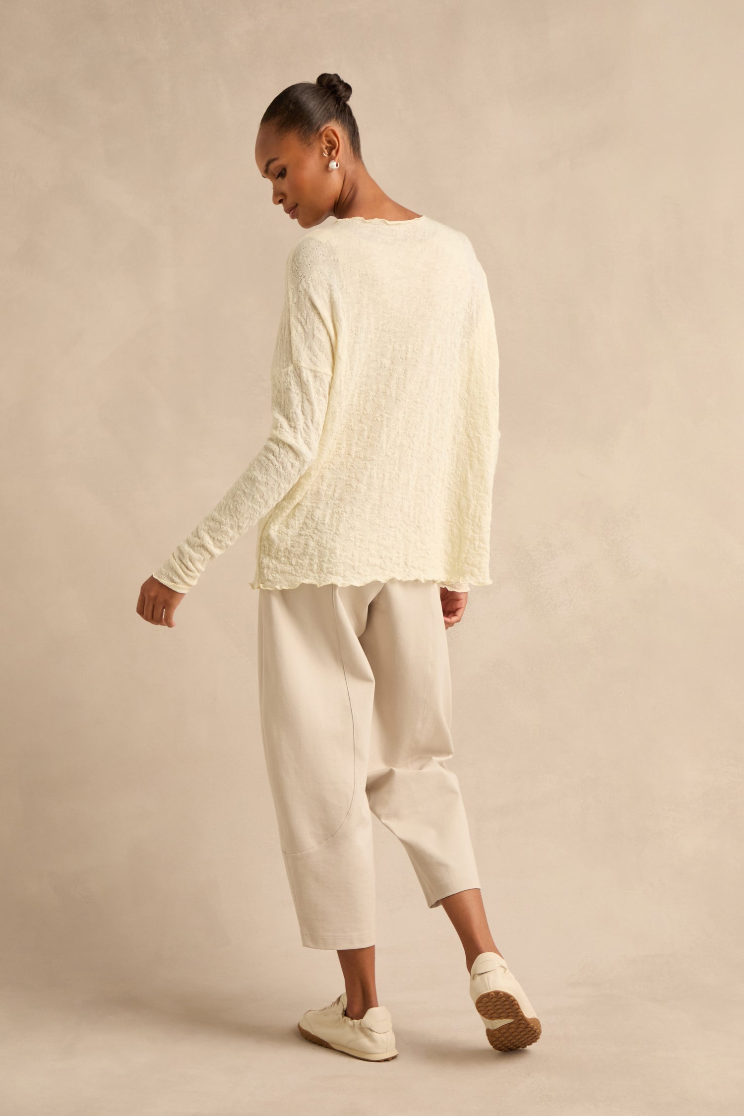 A person stands sideways against a beige studio background, wearing the Ivory Textured Merino Scoop Neck Top, loose light beige pants, and cream slip-on shoes.