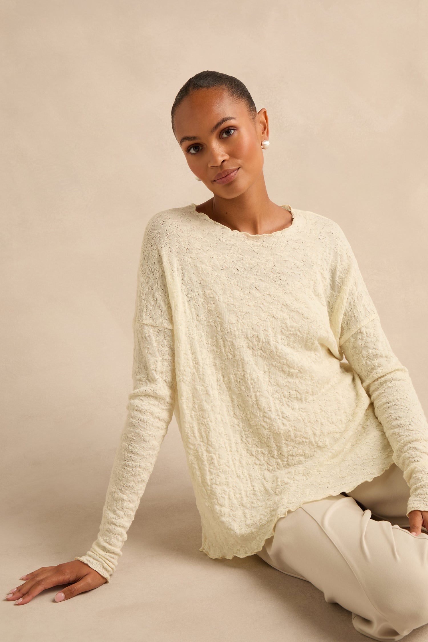 An individual with short hair sits on the floor against a beige background, wearing the Ivory Textured Merino Scoop Neck Top paired with light pants.