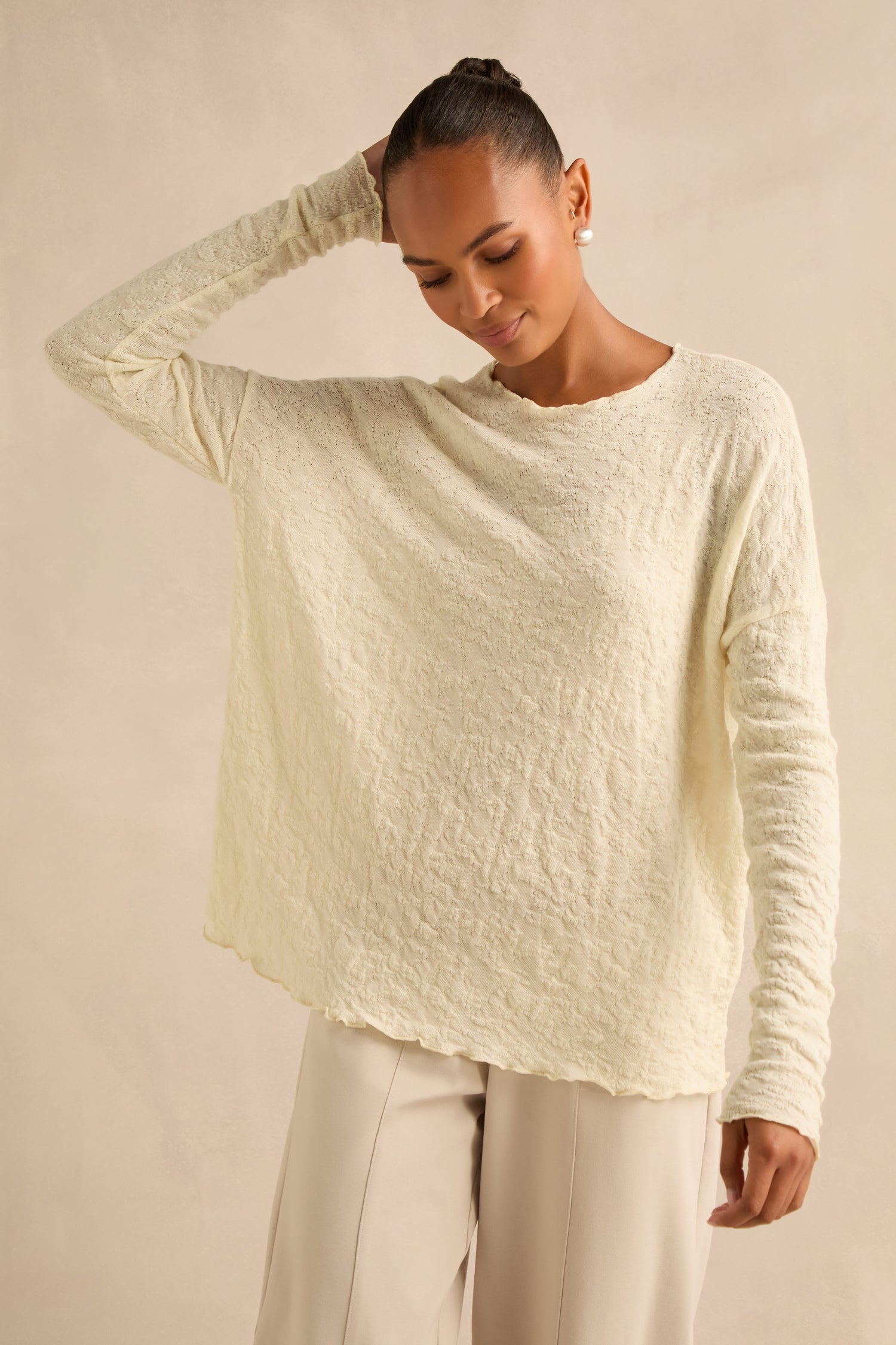 A woman stands against a neutral background, one hand touching her head, wearing the Ivory Textured Merino Scoop Neck Top and light beige pants.