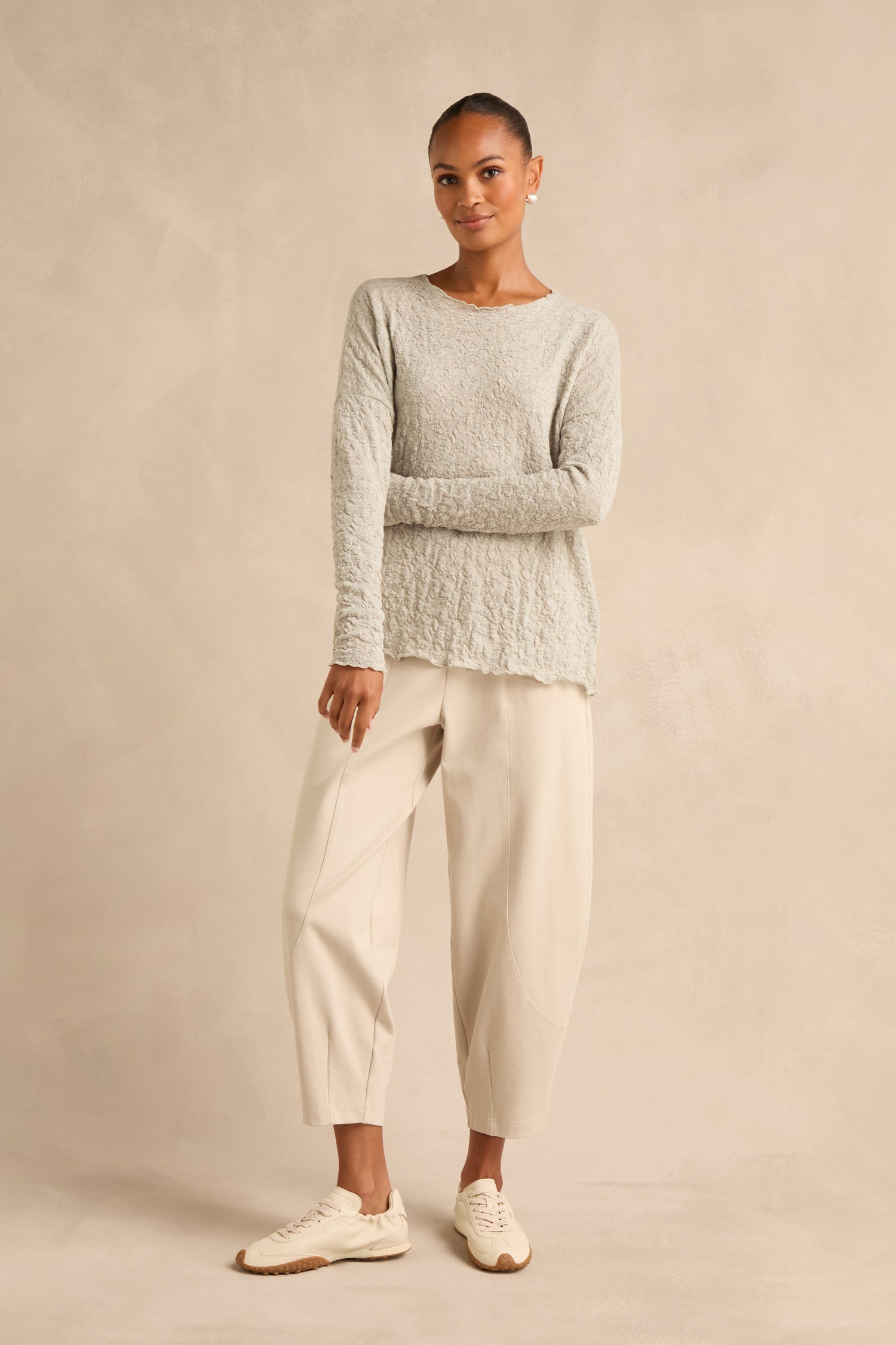 A woman stands against a beige backdrop wearing the Silver Textured Merino Scoop Neck Top, paired with loose cream-colored pants and beige sneakers, her hair pulled back.