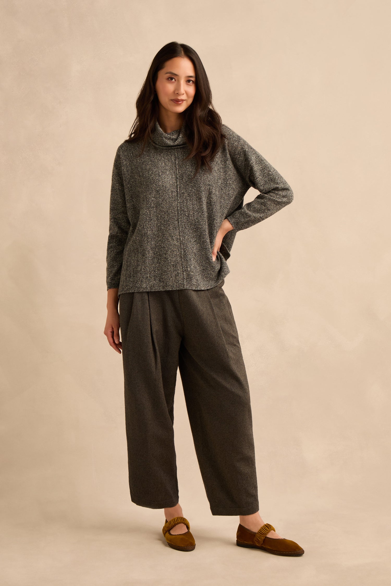 A woman stands against a beige background, wearing the Black & White Merino Wool Cowl Neck Jumper, paired with loose gray pants and brown loafers, with one hand on her hip.