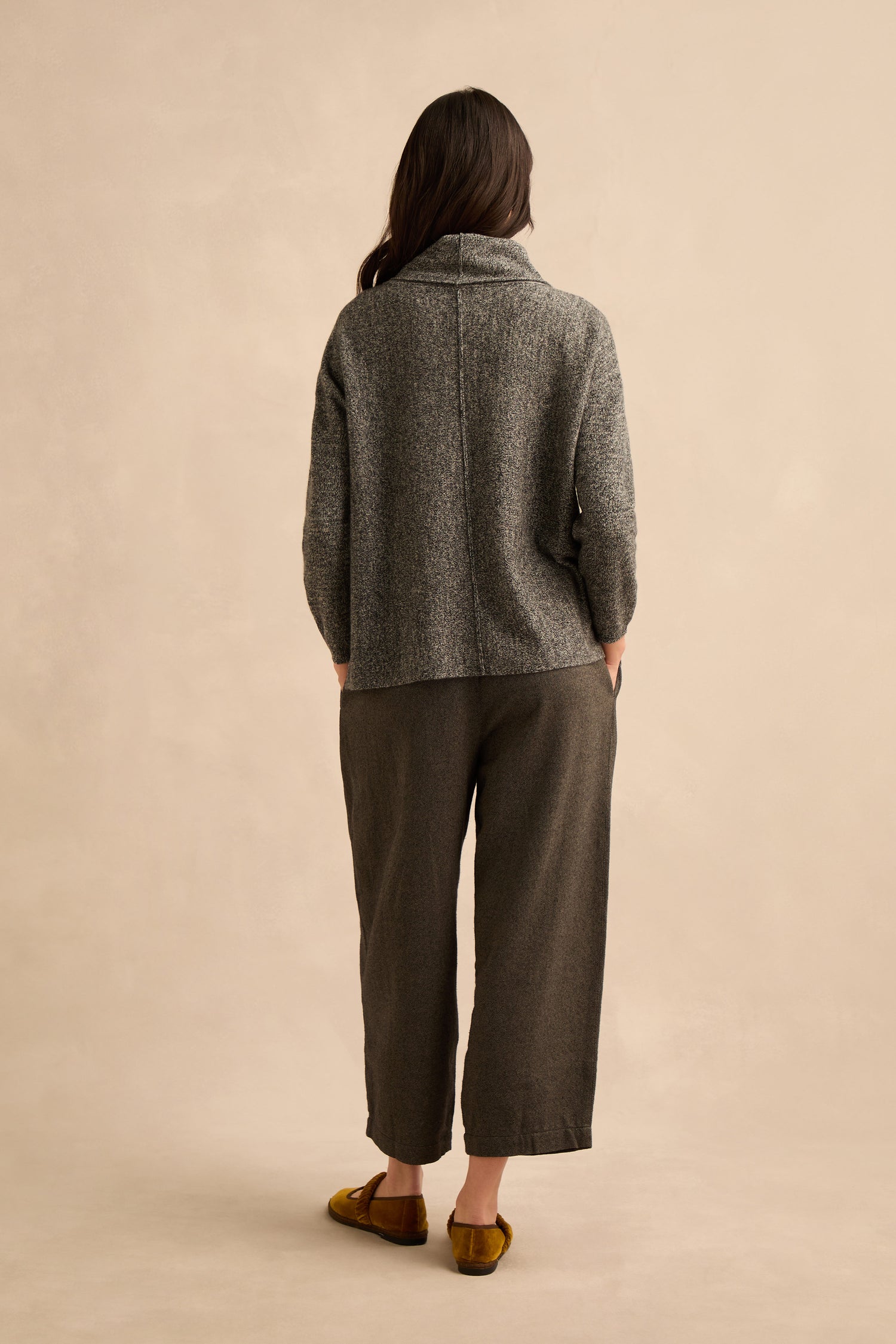 A person is standing with their back to the camera, wearing a cosy Black & White Merino Wool Cowl Neck Jumper, wide-leg dark pants, and yellow slippers, against a neutral background.