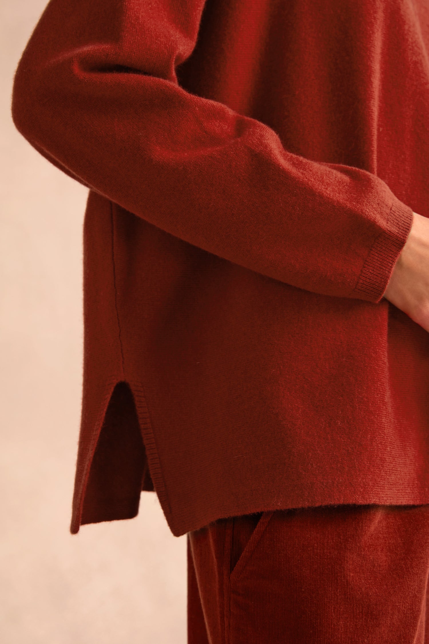 Person wearing the Conker Merino Wool Cowl Neck Jumper, a cosy rust-colored knit with long sleeves and side slits, styled with matching pants and visible from shoulder to hip against a neutral background.