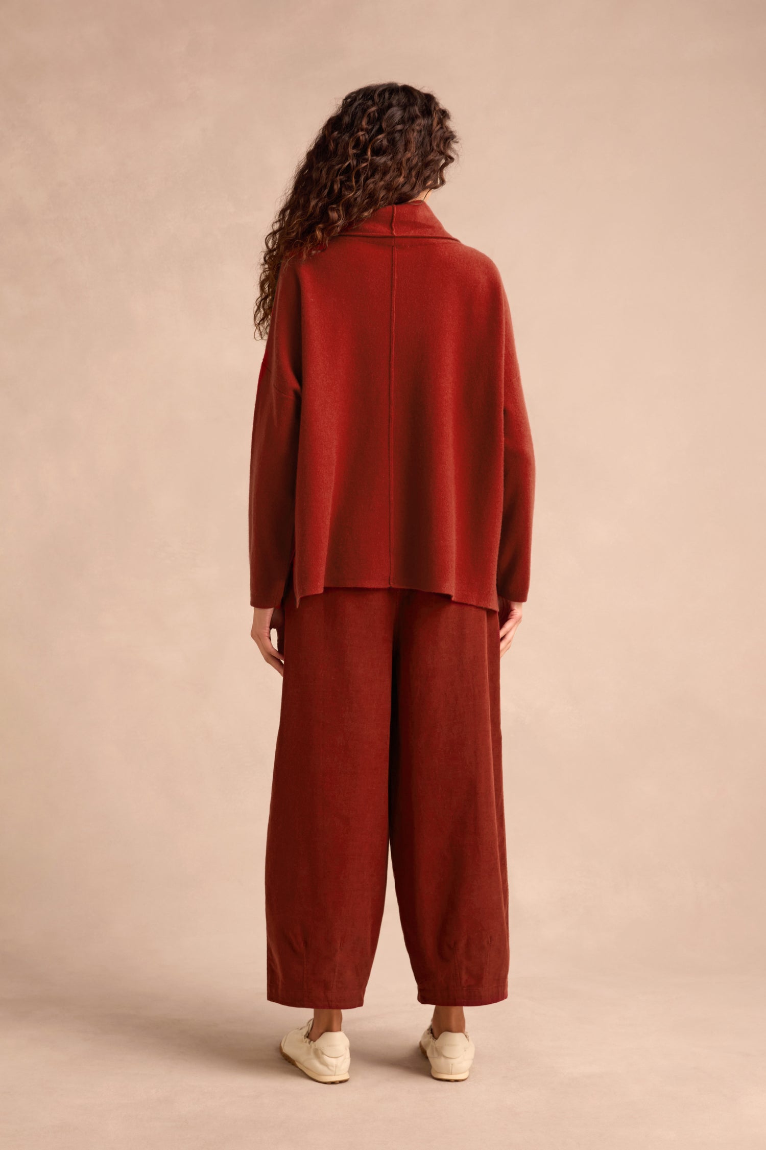 A person stands facing away, wearing a loose-fitting Conker Merino Wool Cowl Neck Jumper in rust and matching wide-leg pants with beige shoes against a neutral background.