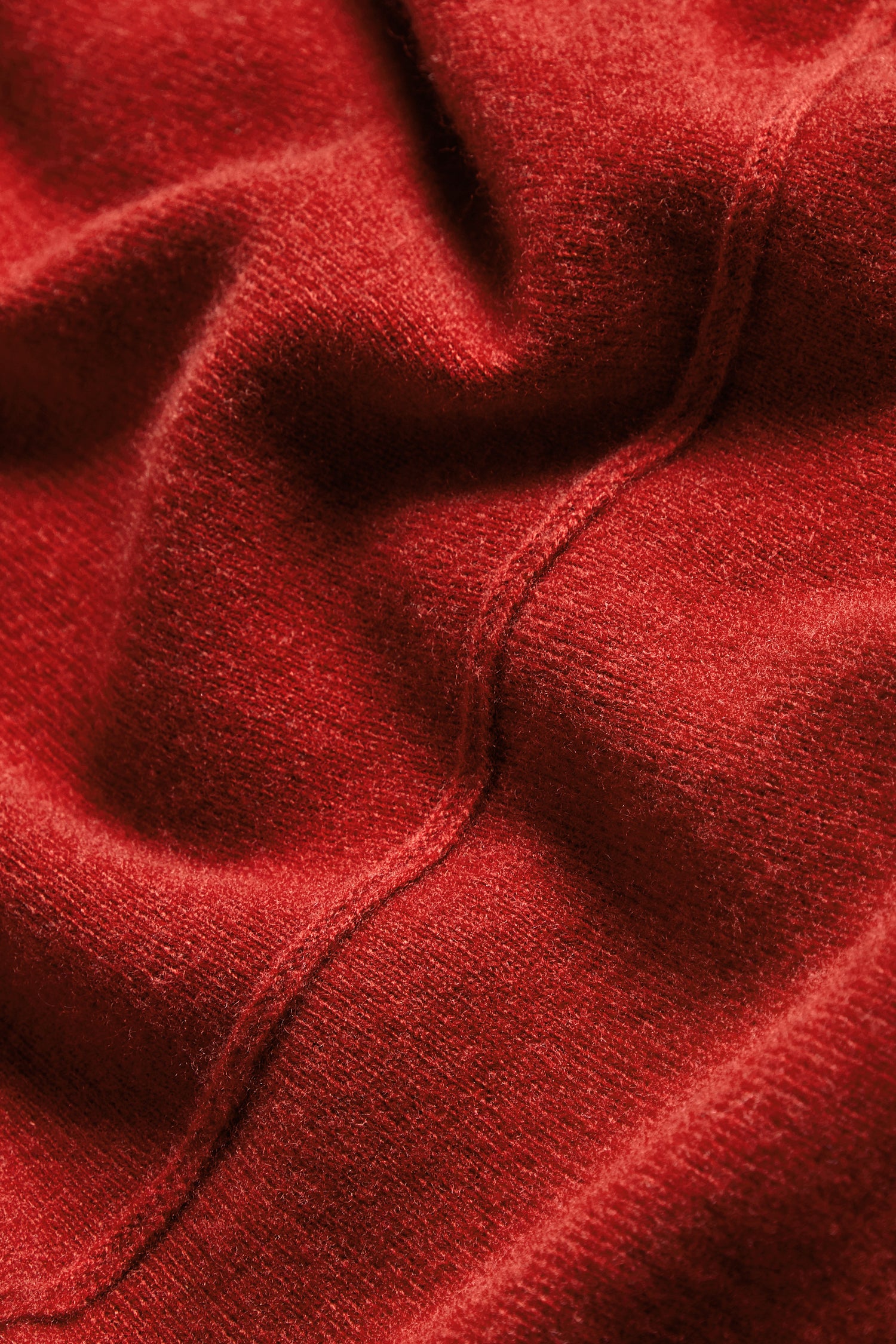 Close-up of the Conker Merino Wool Cowl Neck Jumper reveals its textured red knit with visible seams and gentle folds, highlighting the soft, slightly fuzzy surface—perfect for cosy knitwear.