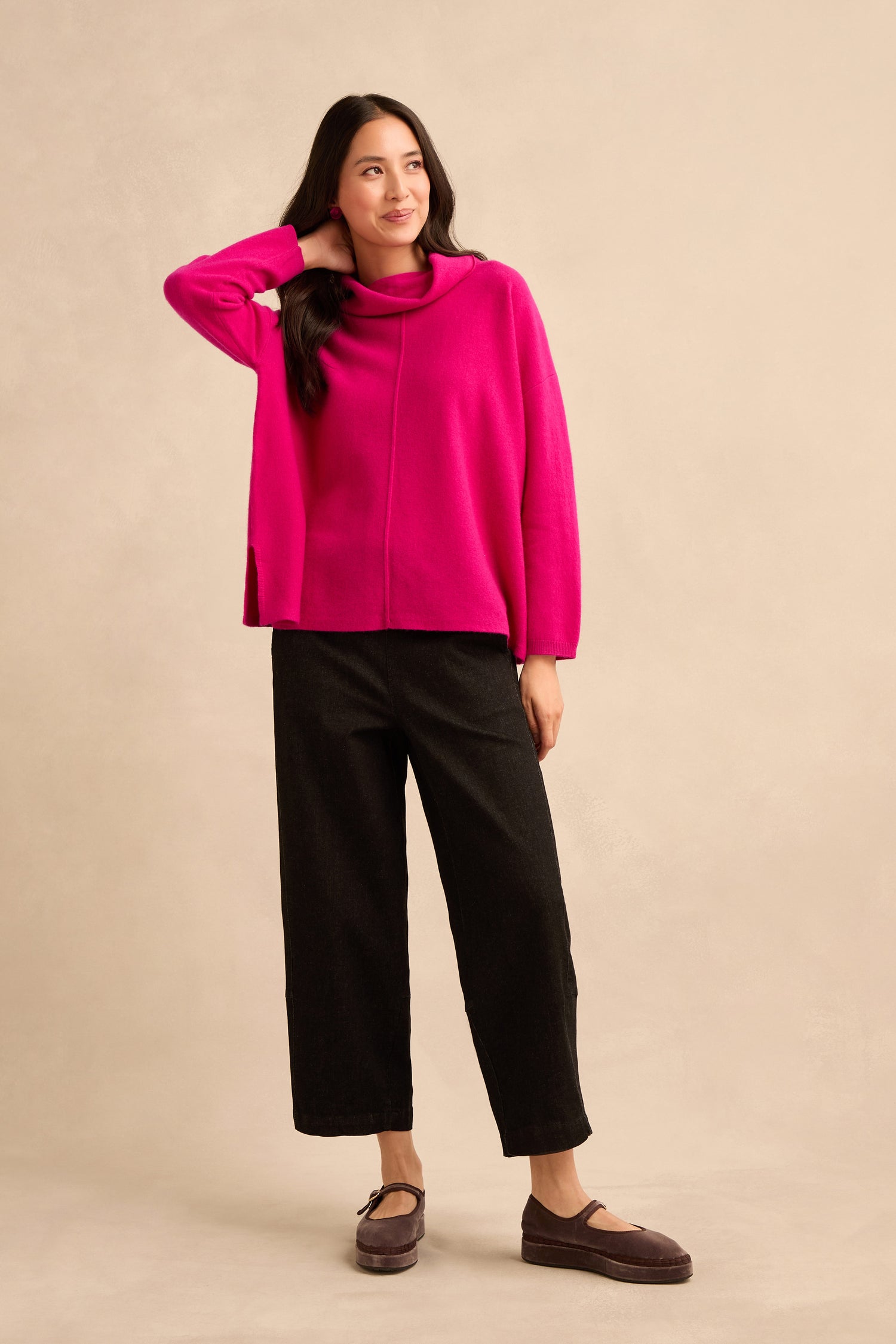 A woman stands against a beige background wearing the Magenta Merino Wool Cowl Neck Jumper, paired with black wide-leg pants and brown shoes, with one hand touching her hair.