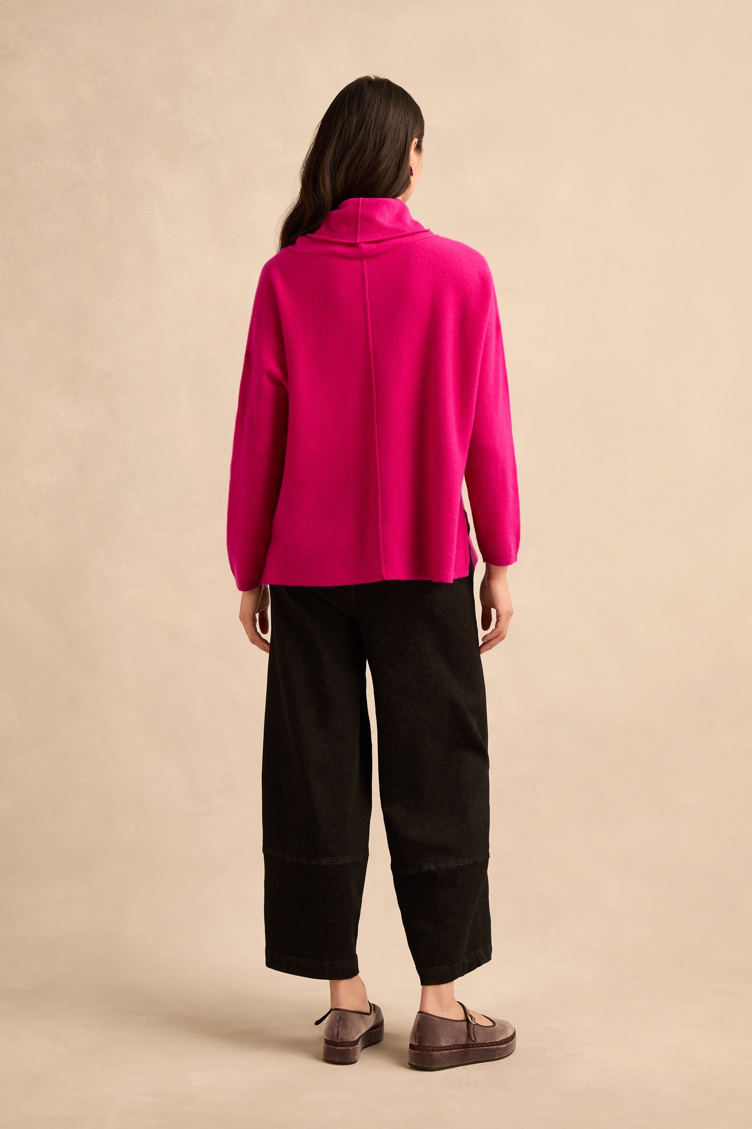 A person stands facing away, wearing a Magenta Merino Wool Cowl Neck Jumper, loose black pants, and brown shoes against a beige background.