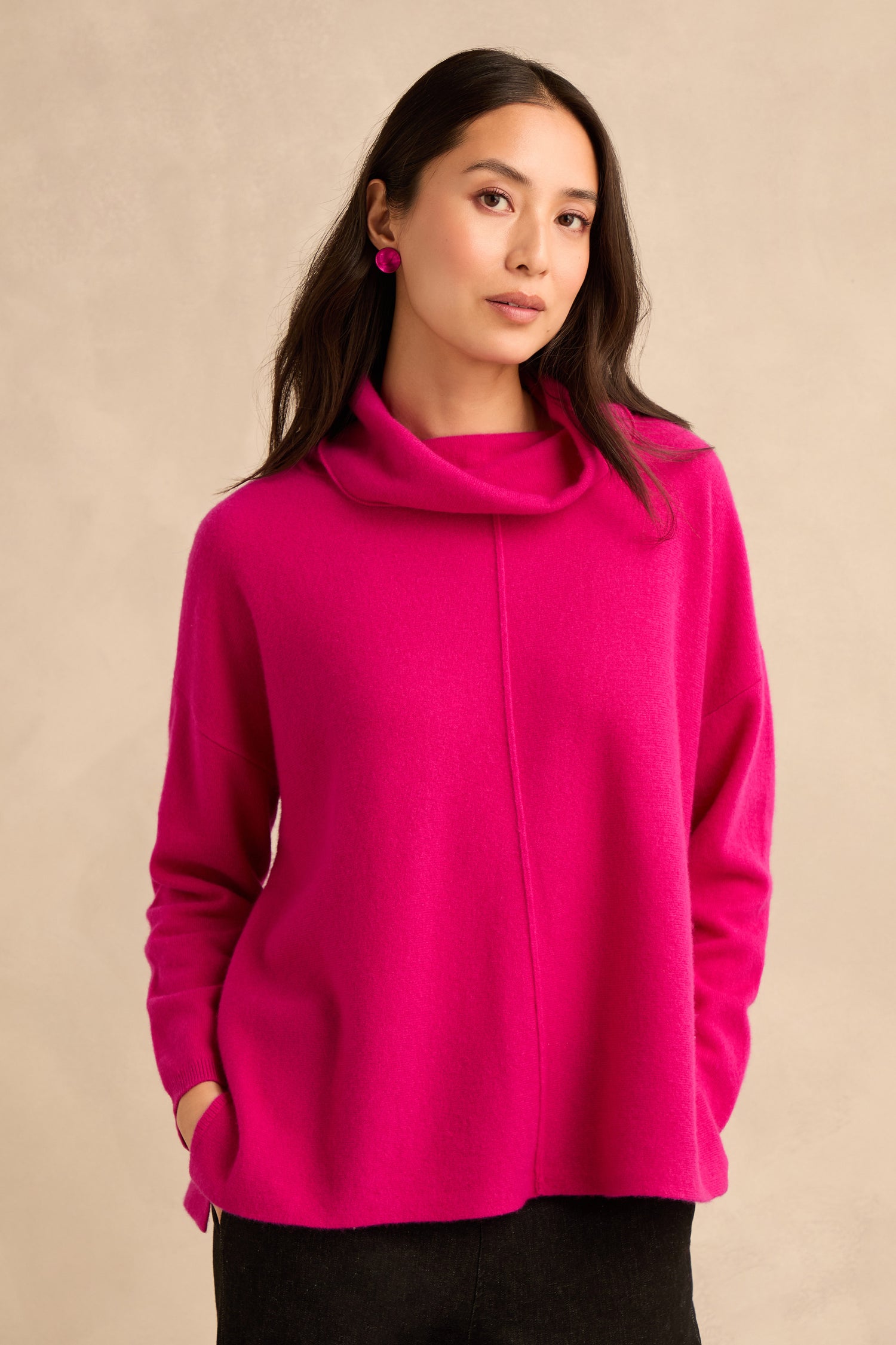 A woman wearing the Magenta Merino Wool Cowl Neck Jumper, featuring a centre seam detail, styled with black trousers and matching pink earrings—the perfect cosy knit sweater look for cooler days.