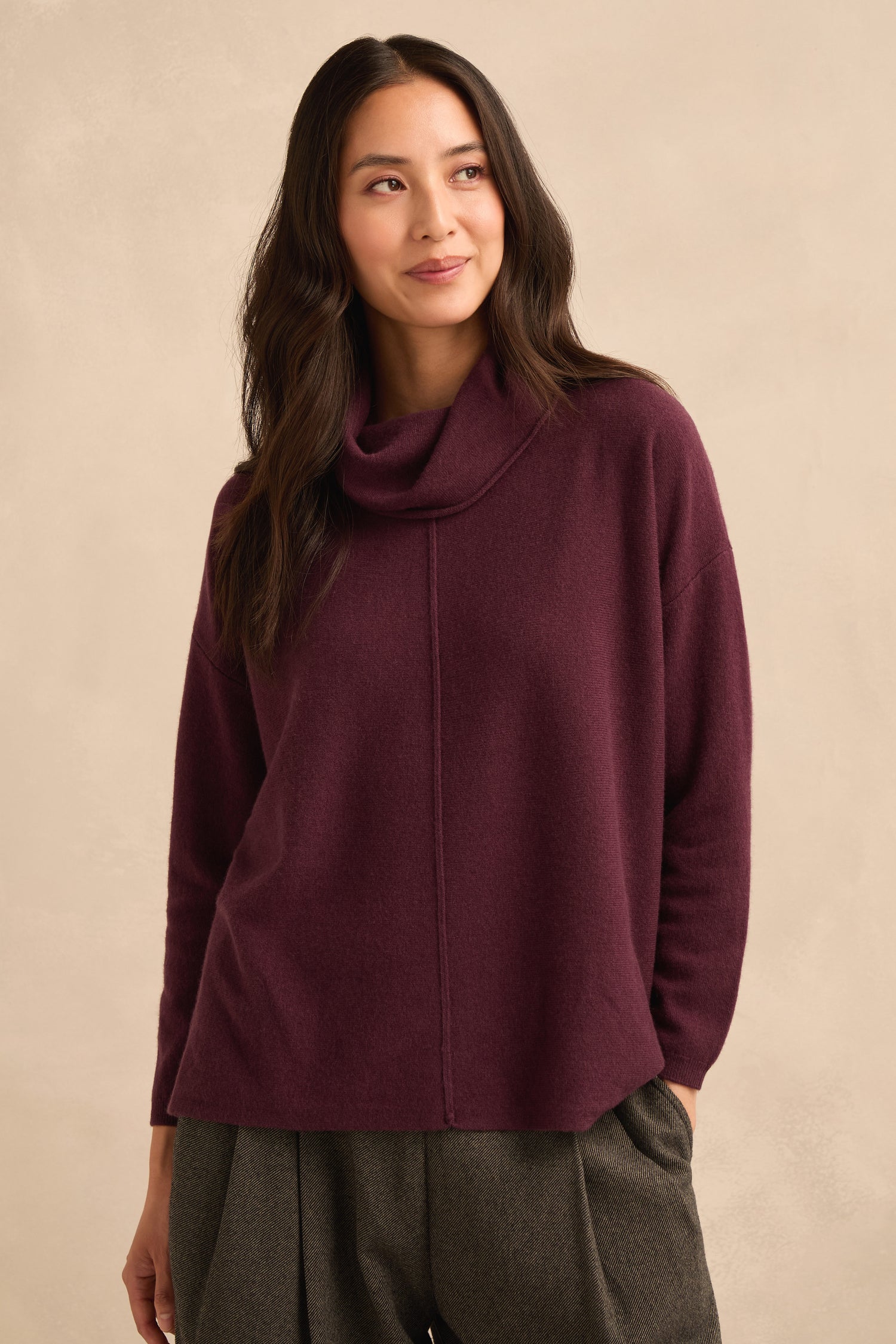 A woman wearing a Plum Merino Wool Cowl Neck Jumper and gray trousers stands against a beige background, gazing slightly to the side.