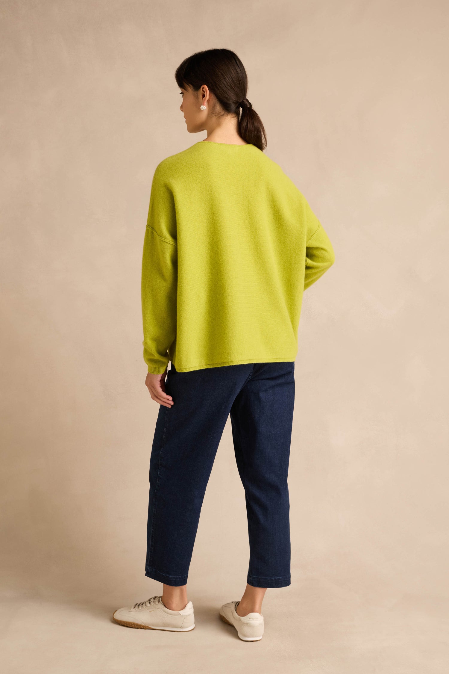 A person stands with their back to the camera, wearing the Kiwi Merino Wool V Neck Jumper in lime green, paired with dark blue jeans and white sneakers, set against a plain beige background.
