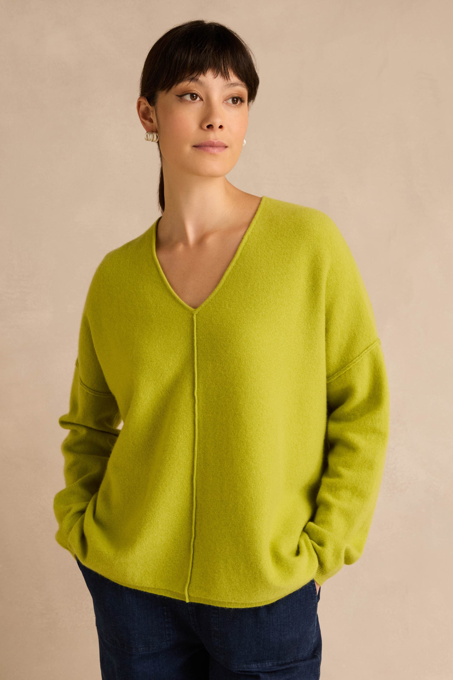 A woman wearing the Kiwi Merino Wool V Neck Jumper in lime green and dark blue pants stands against a beige background with her hands in her pockets.