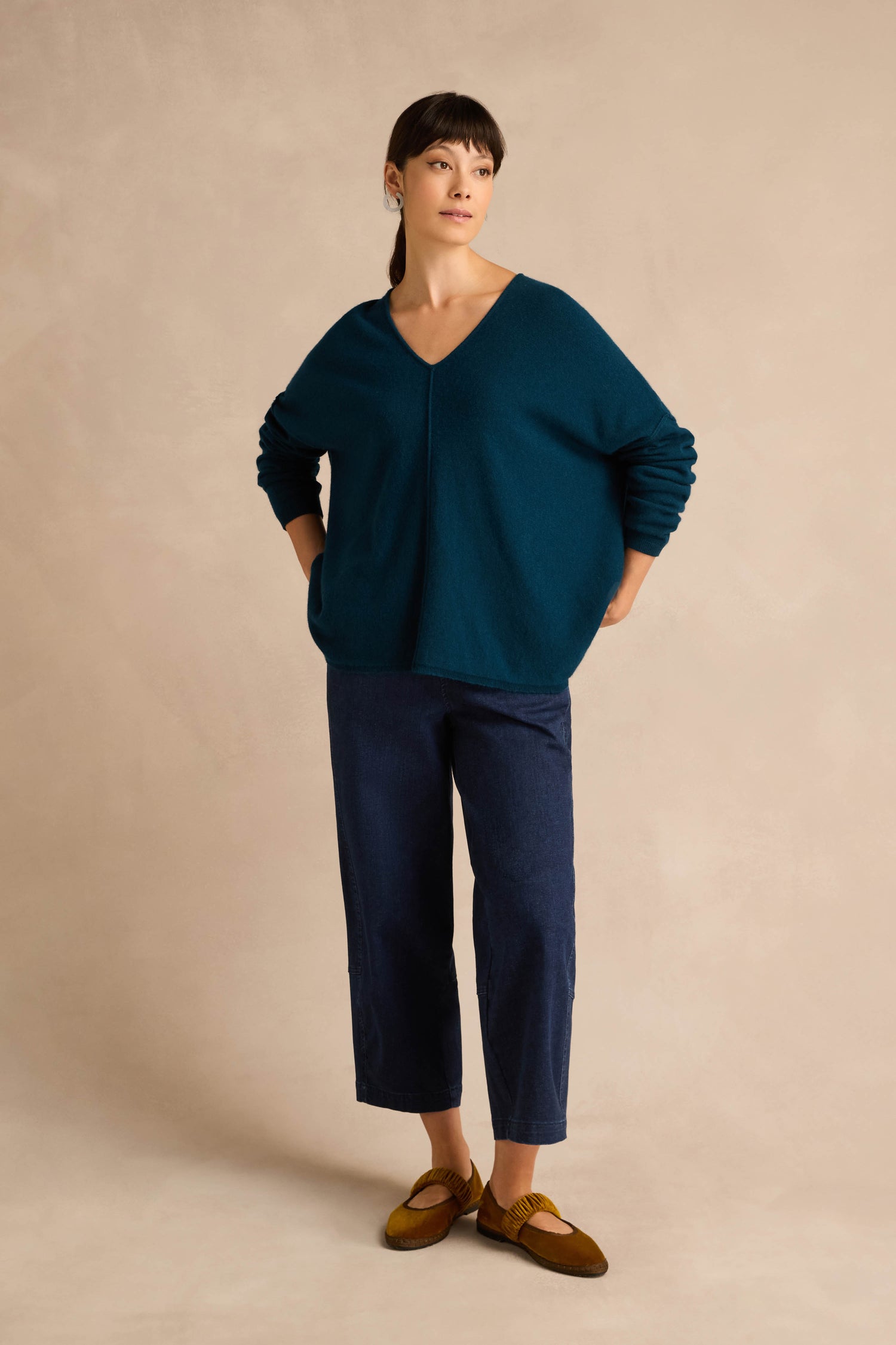 A woman stands against a beige background, wearing the Marine Merino Wool V Neck Jumper in teal, paired with dark blue cropped pants and brown shoes, with her hands in her pockets.