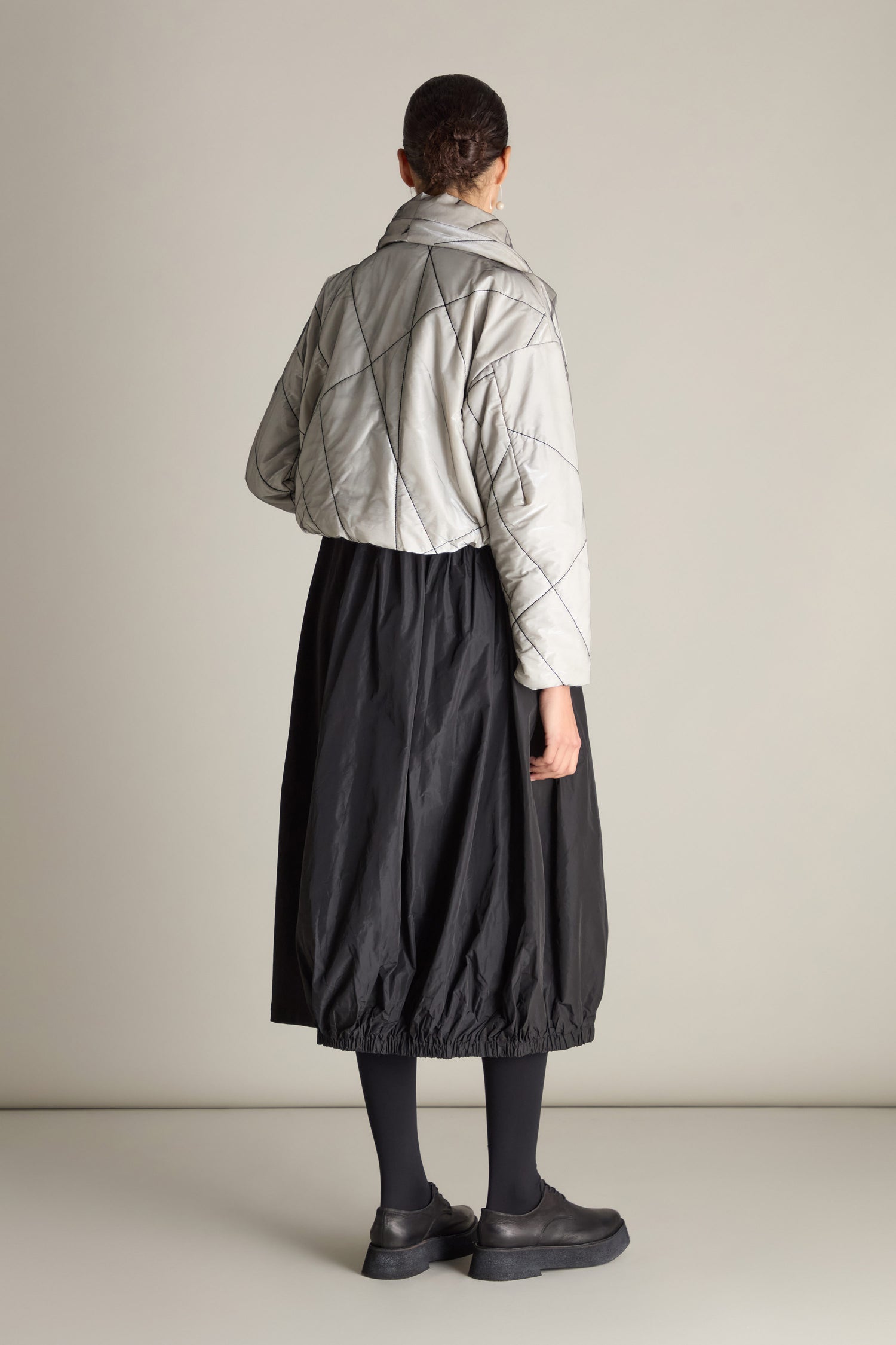 A person is seen from the back wearing a Short Puffa Evening Jacket with embroidered detailing, paired with a black skirt, black tights, and black shoes. The background is plain and neutral.