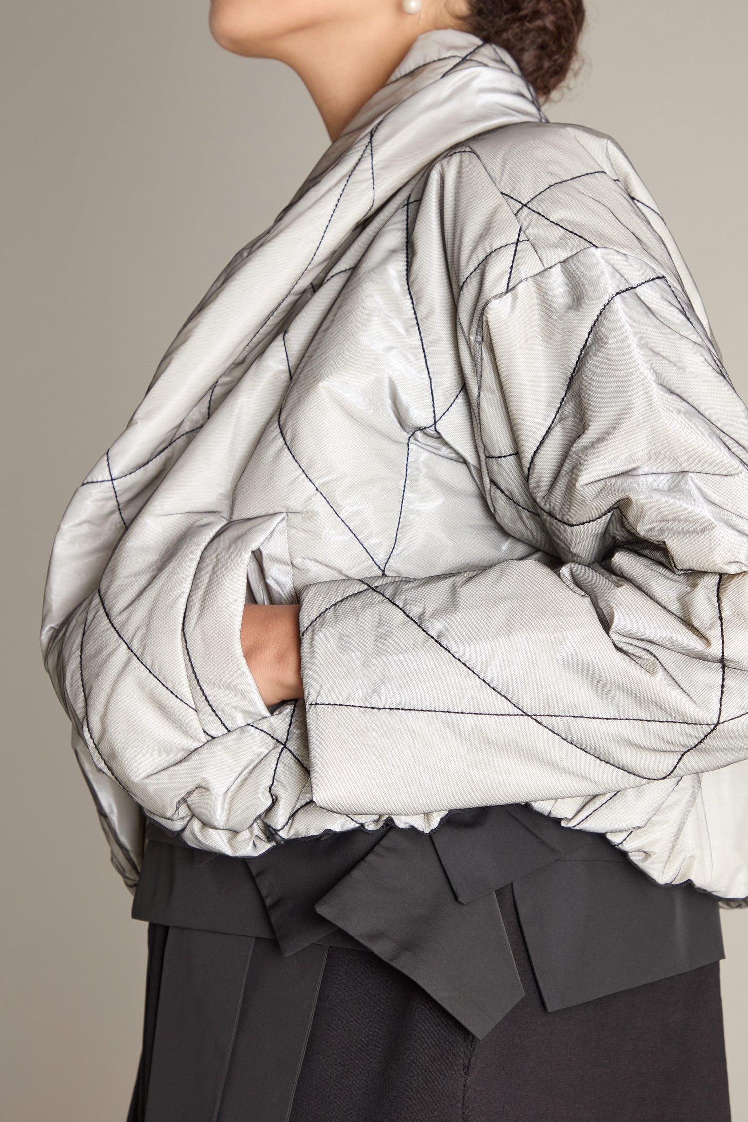 A person is shown in profile wearing a Short Puffa Evening Jacket, which features a silver, geometric pattern. Their hands are tucked into the pockets of the jacket, and their head is tilted slightly upwards. They are also dressed in dark pants.