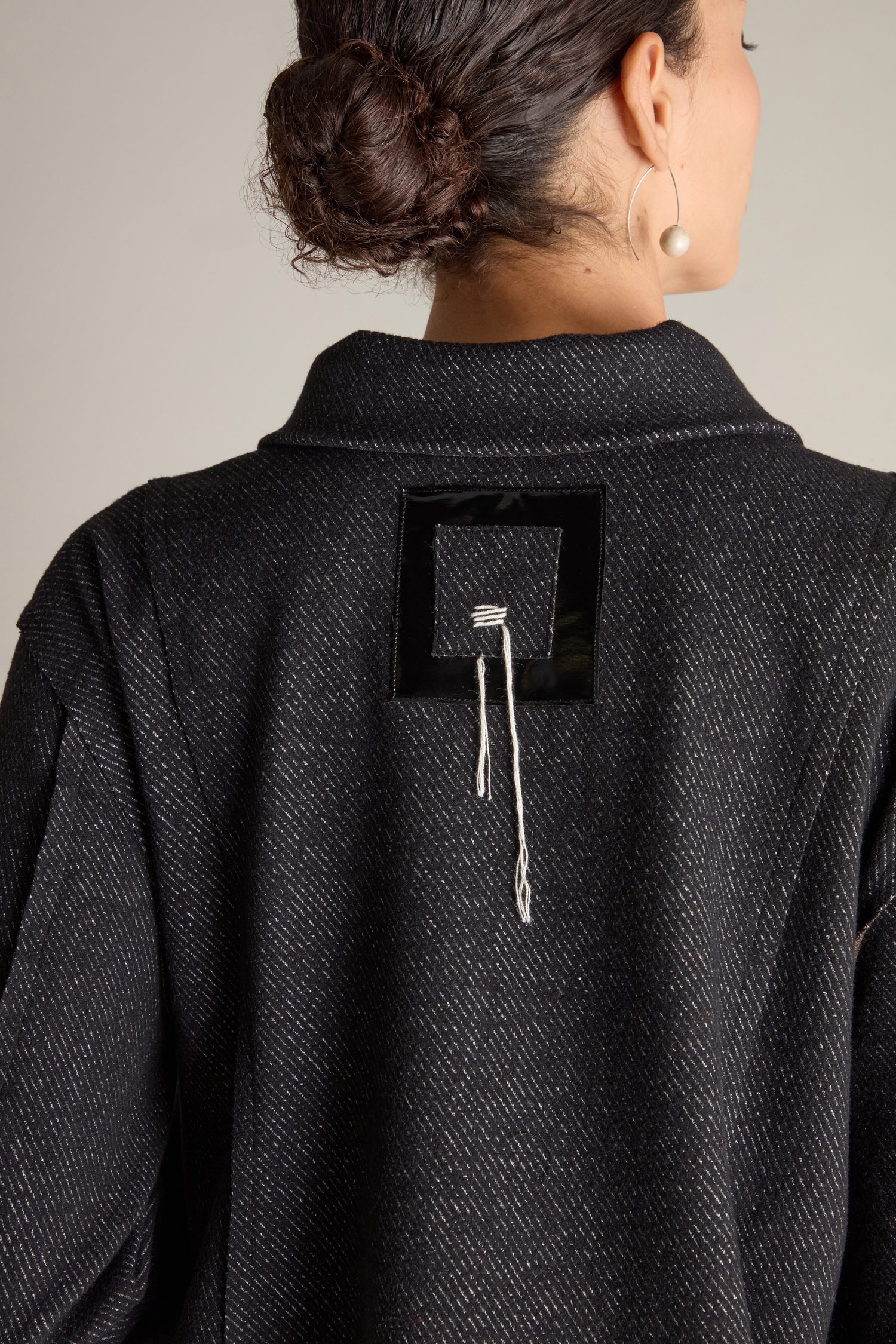 A person is seen from behind wearing the Zip Detail Jacket, which features a square patch and dangling strings on the back. Their hair is tied up, showcasing a small hoop earring with a pearl. The jacket's oversized front pockets contribute to its stylish appeal.