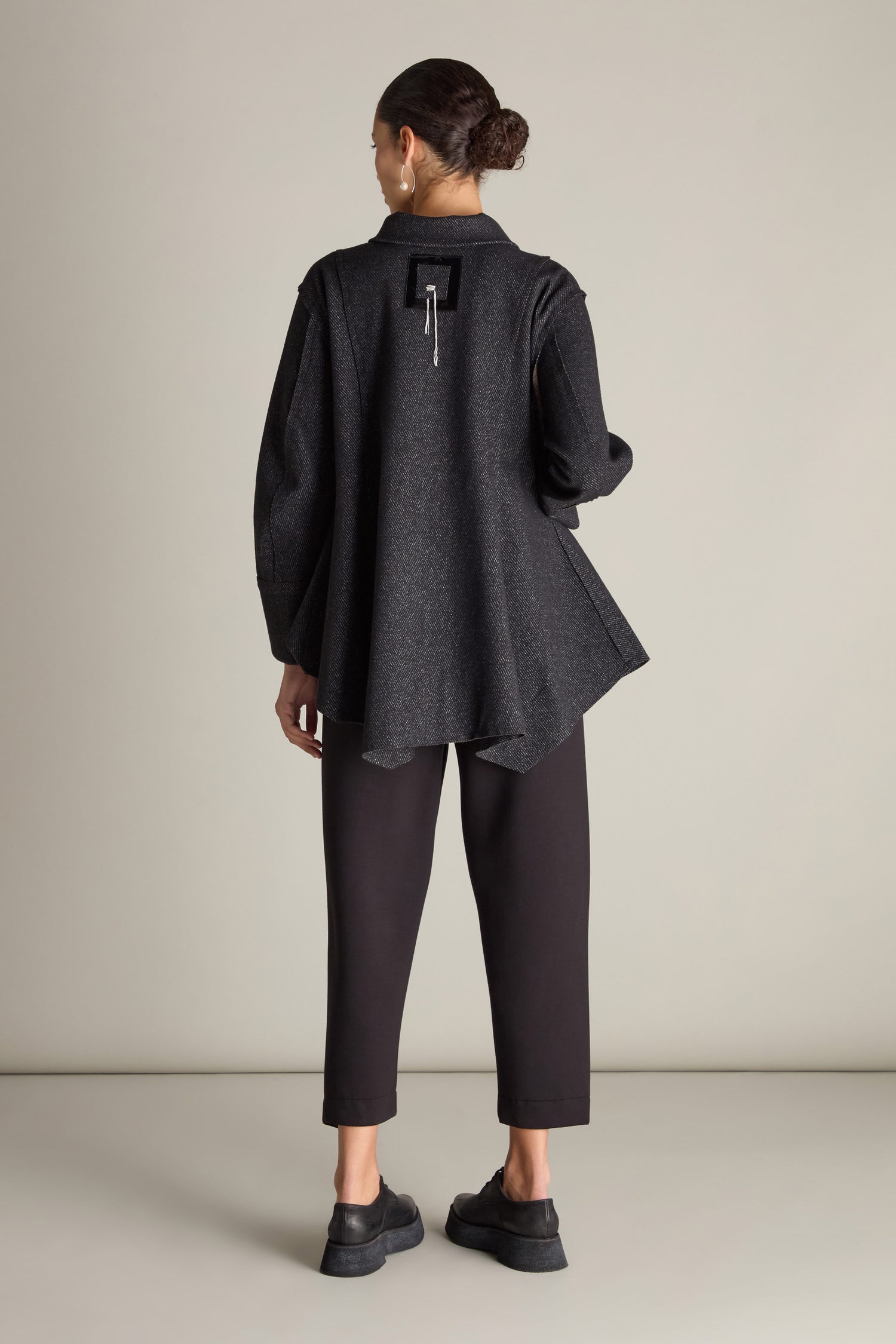 A person with dark hair tied back is seen from the back, wearing a Zip Detail Jacket in long dark grey with an asymmetrical hem and oversized front pockets, black pants, and black shoes, standing against a plain background.