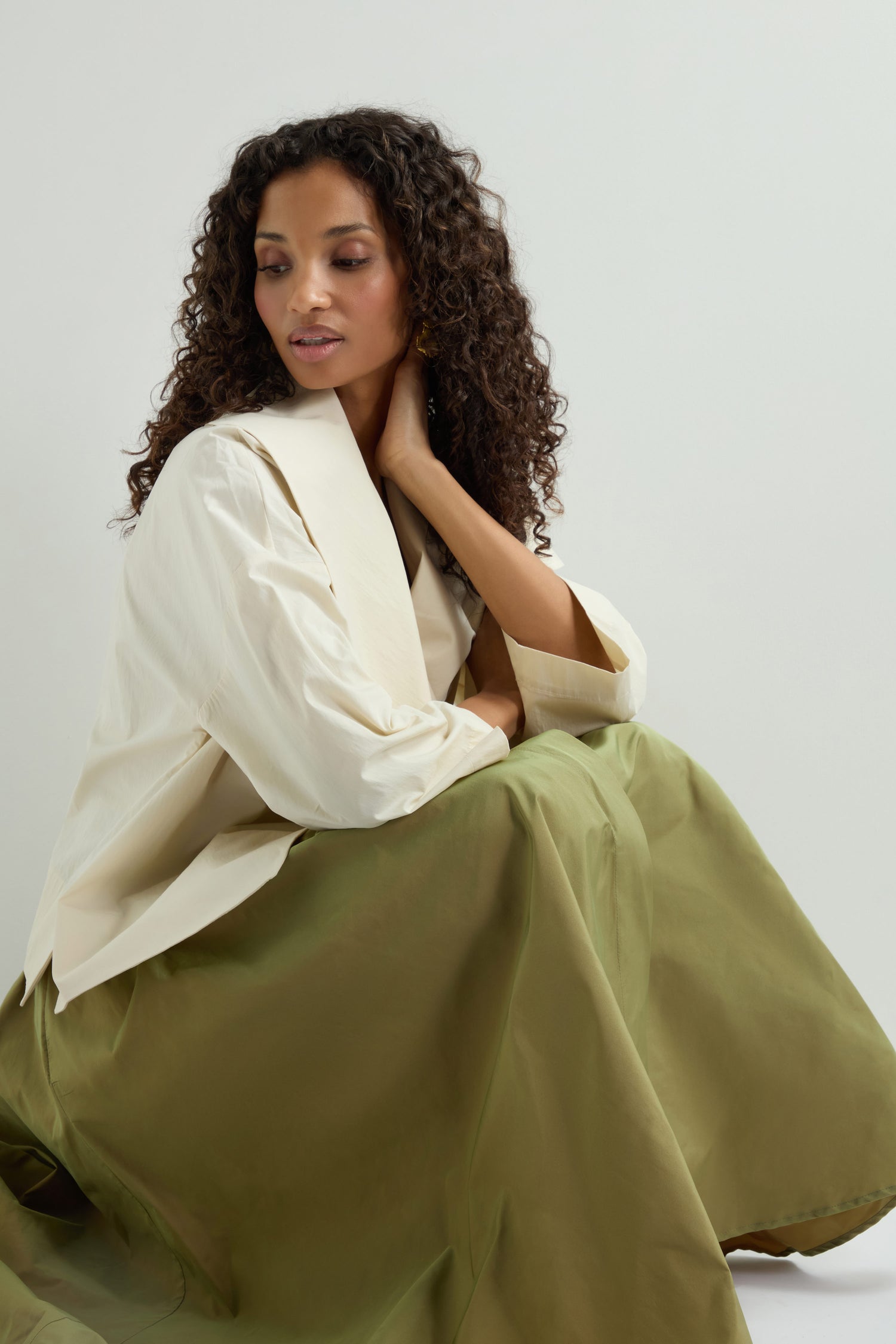 A person with long curly hair, wearing a white top and a green Taffeta Skirt, is sitting with one hand on their neck against a plain backdrop.