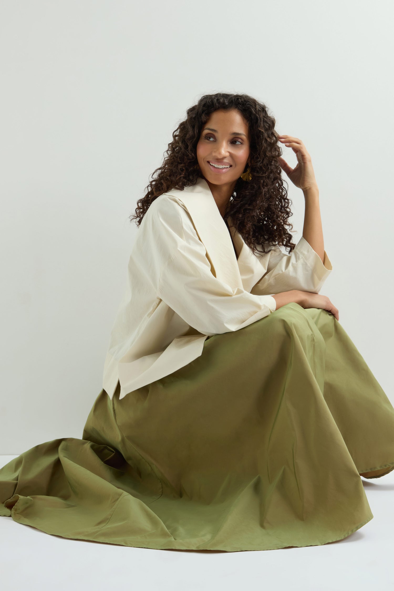 A person with curly hair smiles, wearing a light beige top and an elegant A-line long olive green Taffeta Skirt, sitting against a plain backdrop.