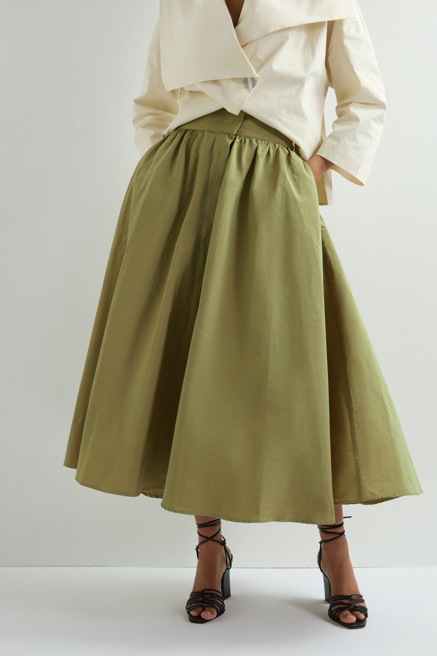 A person models a green Taffeta Skirt—a midi A-line design with pockets—along with an off-white top. They complete the look elegantly with black strappy heels against a light background.