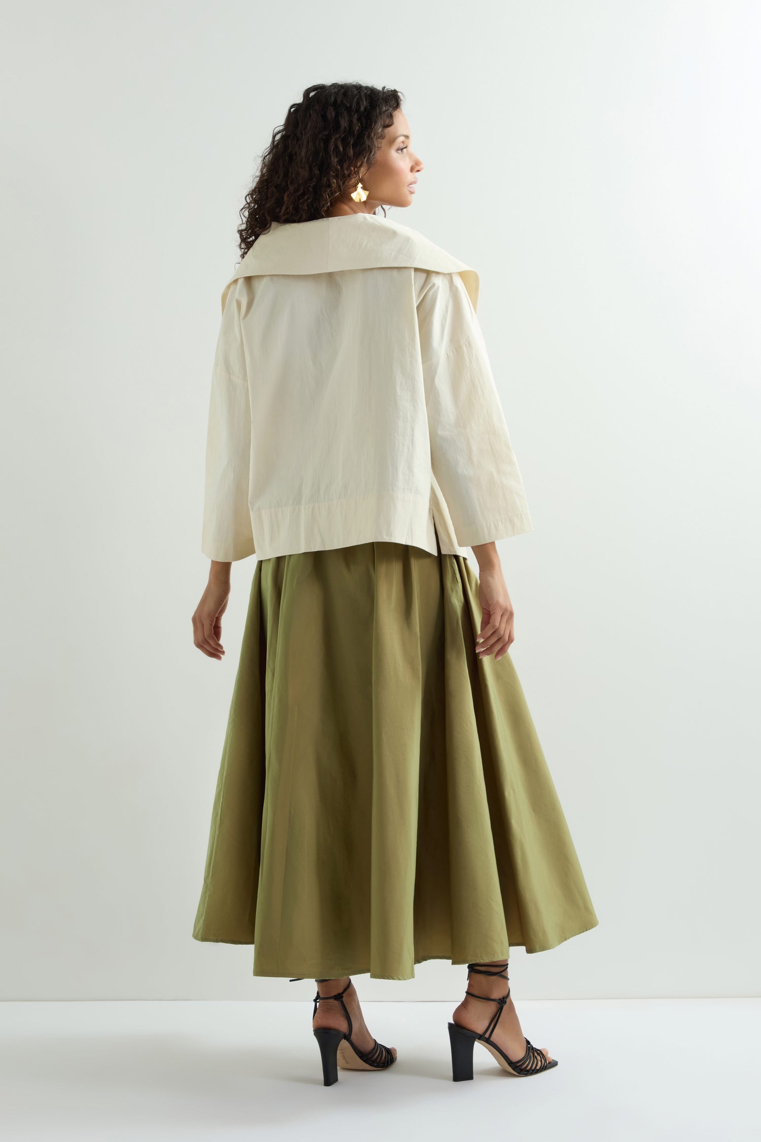 A woman faces right, wearing a light-colored jacket with an olive-green Taffeta Skirt in A-line midi-length style. She completes her look with black high-heeled sandals, all set against a crisp white backdrop.
