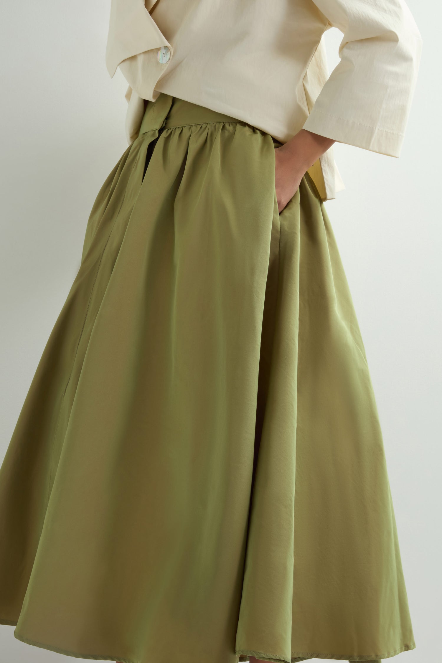 A person wearing a light-colored blazer and an olive green A-line Taffeta Skirt with hands in pockets, set against a neutral background.