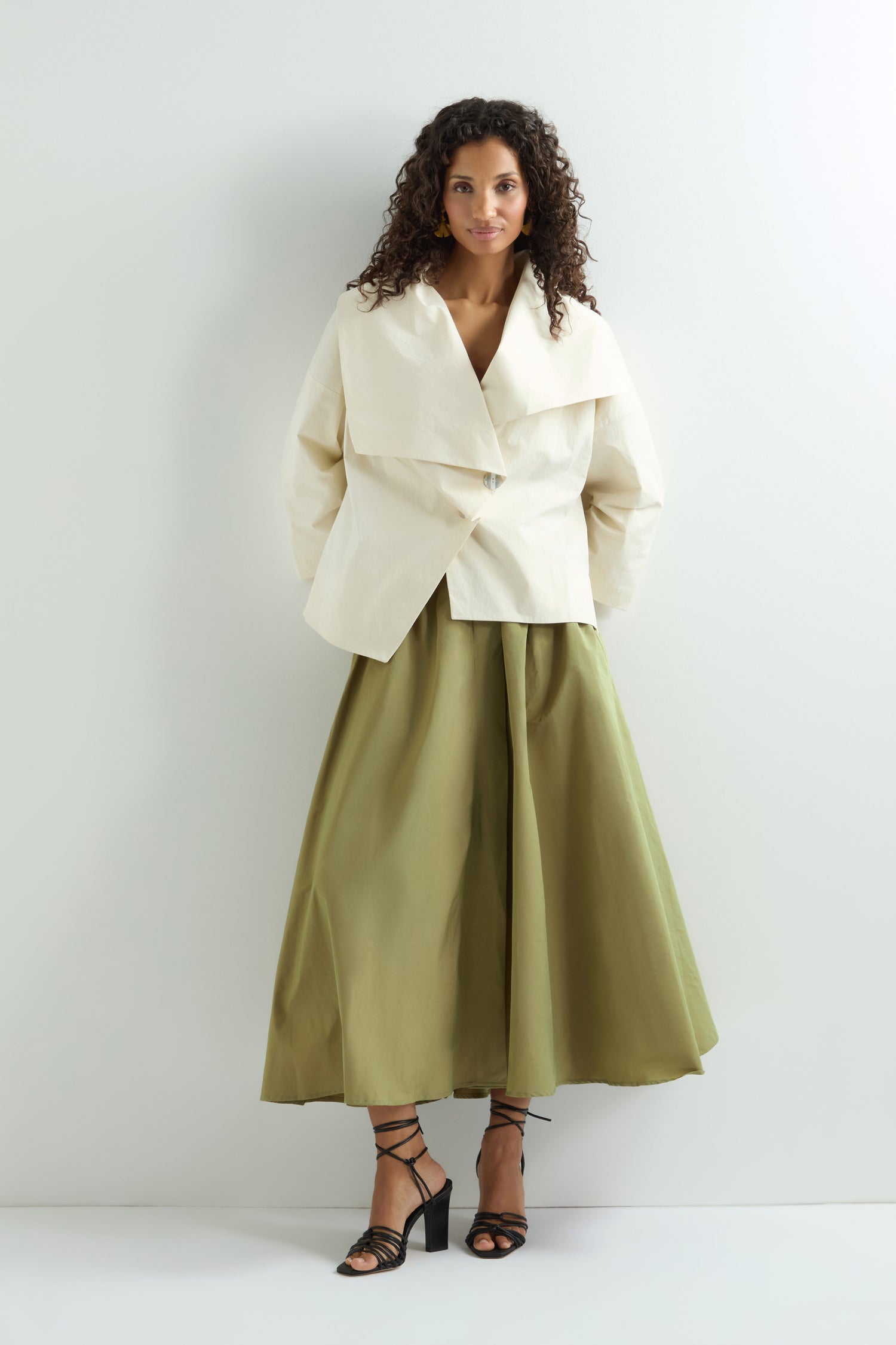 A woman with curly hair models a white jacket and a Taffeta Skirt, midi length and A-line style, paired with black strappy heels against a plain backdrop.