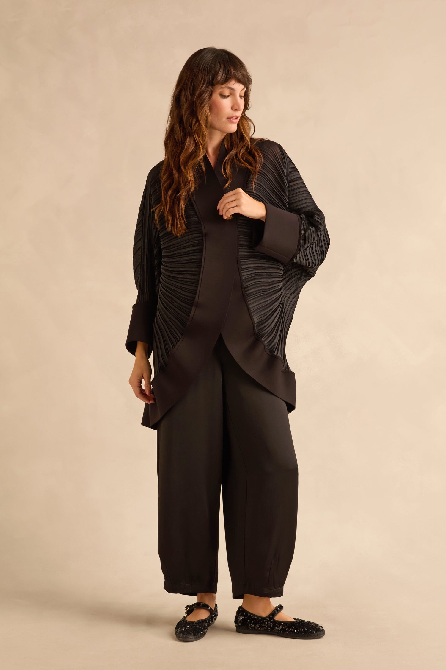 A person wearing a black Pleated Jacket with metallic button details and wide-leg trousers.