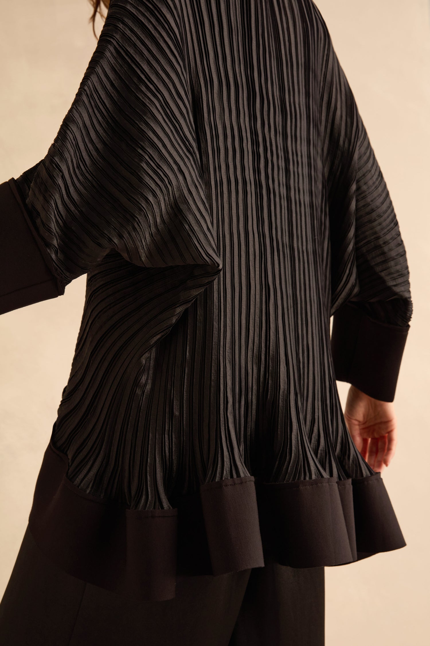 Back view of a black, pleated Pleated Jacket with wide, cuffed batwing sleeves and a flared hem finished with metallic button detail.