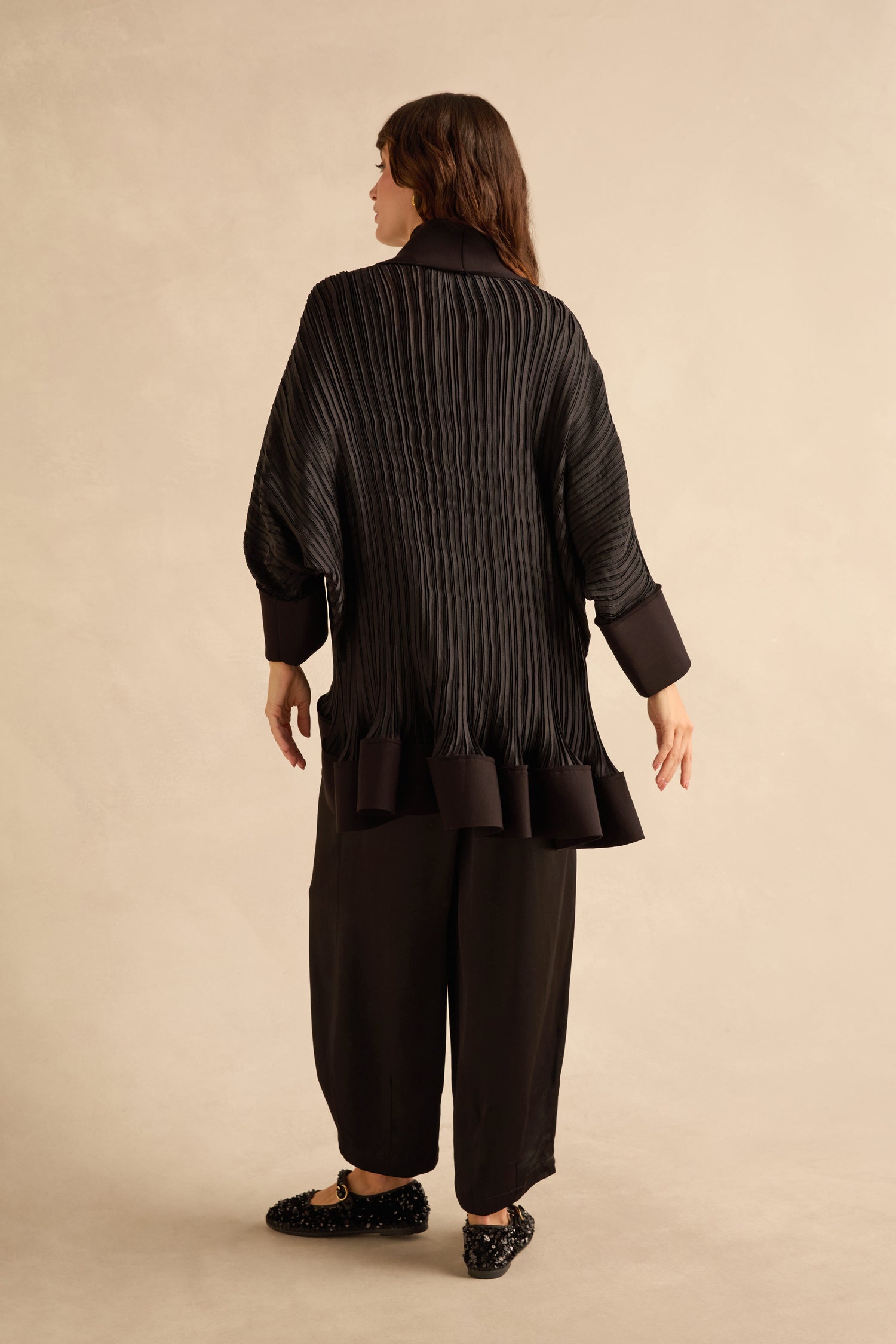 A person seen from behind wears an oversized black Pleated Jacket with batwing sleeves, paired with black trousers and embellished flats.