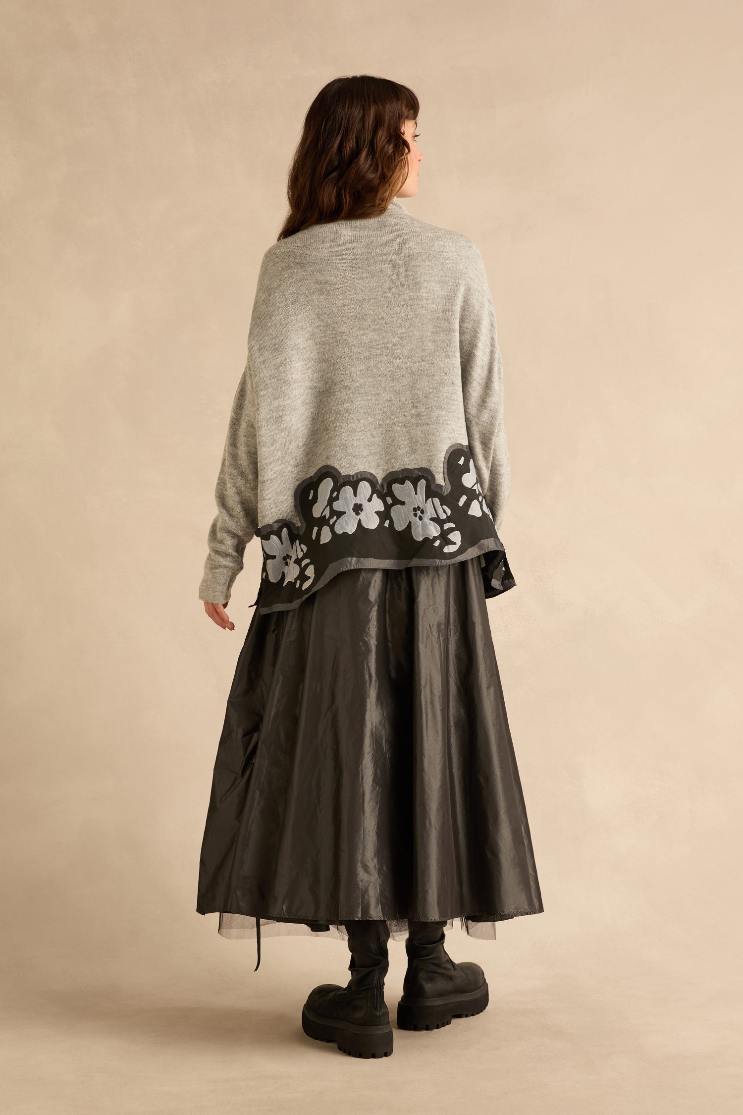 A person seen from the back wearing a grey Flower Hem Knit sweater with floral appliqué, a long black skirt, and black platform boots.
