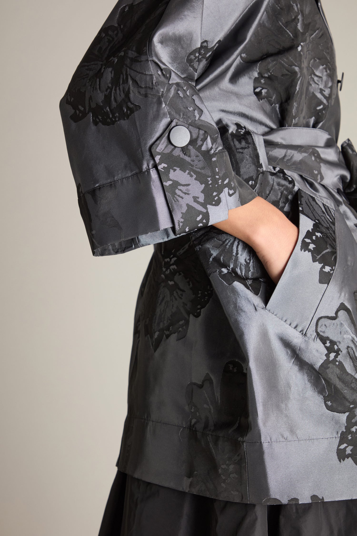 Close-up of a person wearing the Flower Belted Jacket, featuring a shiny, dark-colored trench coat with a floral woven jacquard pattern. The person's hand is in the pocket, and only the torso and part of one arm are visible. The double-breasted button fastening adds an elegant touch to the overall look.