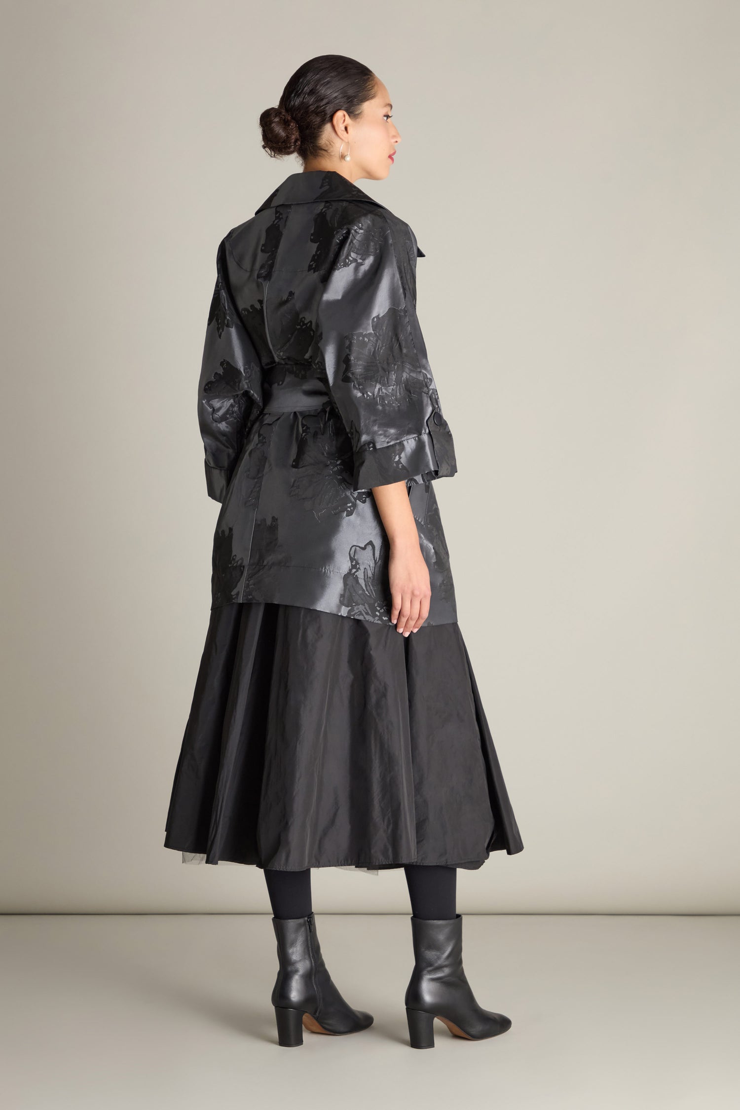 A person with tied-back hair is standing sideways, wearing a long black Flower Belted Jacket with double-breasted button fastening, complemented by a black skirt, tights, and heeled boots in front of a plain background.