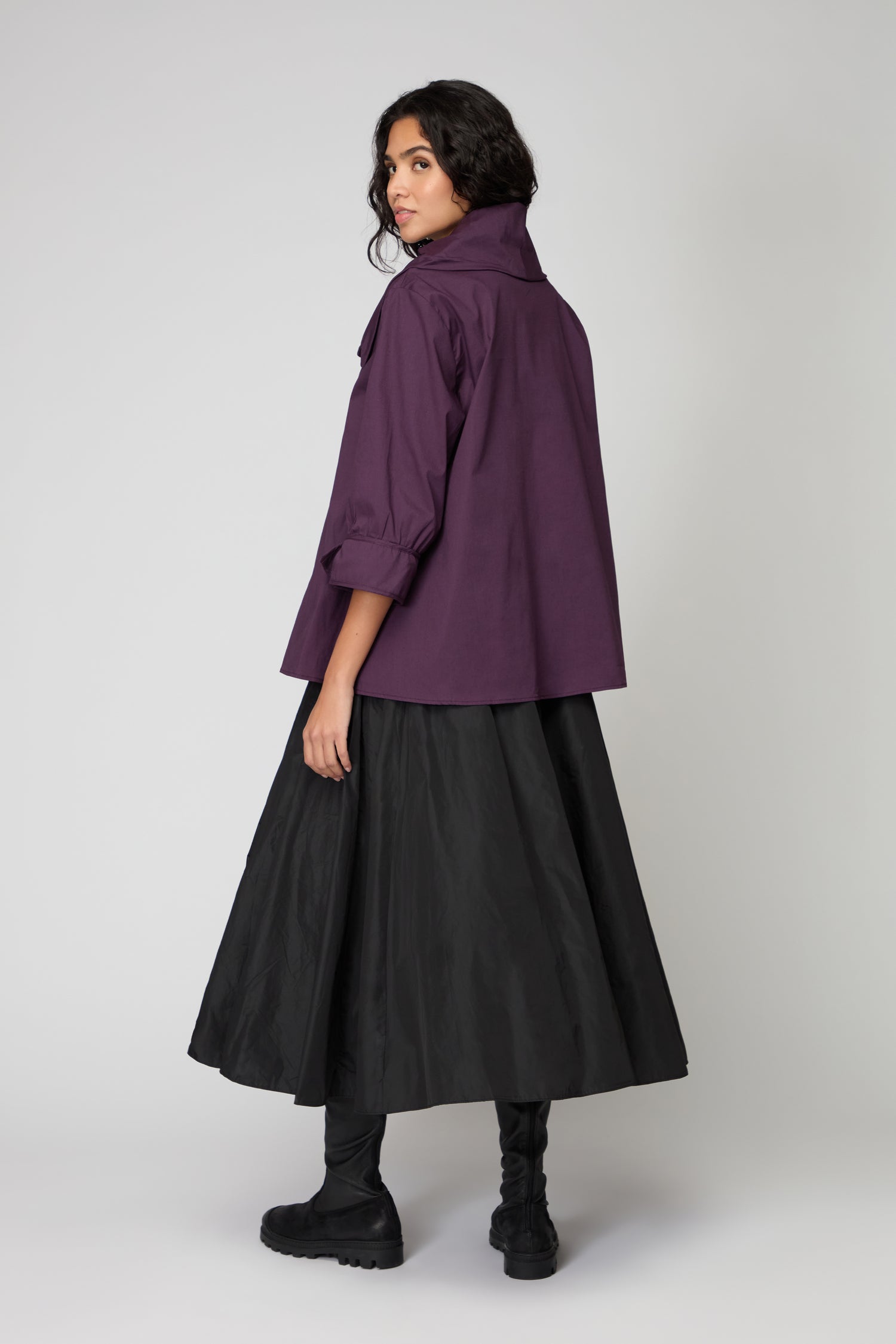 A person faces away, wearing a purple Frill Neck Easy Top with rolled sleeves, a long black skirt, and black boots, set against a plain light background.