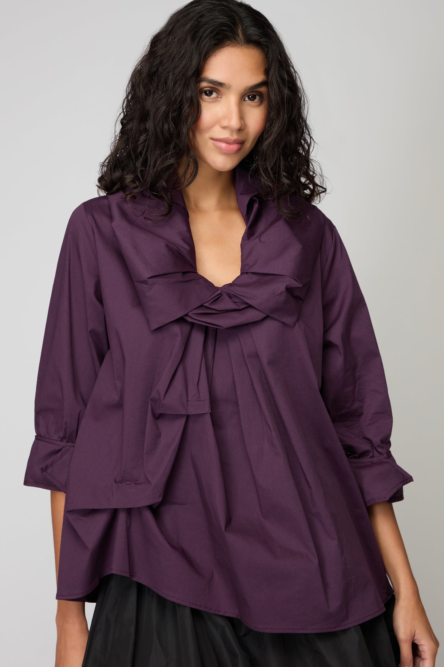 A woman with wavy dark hair wears the Frill Neck Easy Top—a loose-fitting, long-sleeved purple blouse featuring sculptural maximalism with a large front bow.