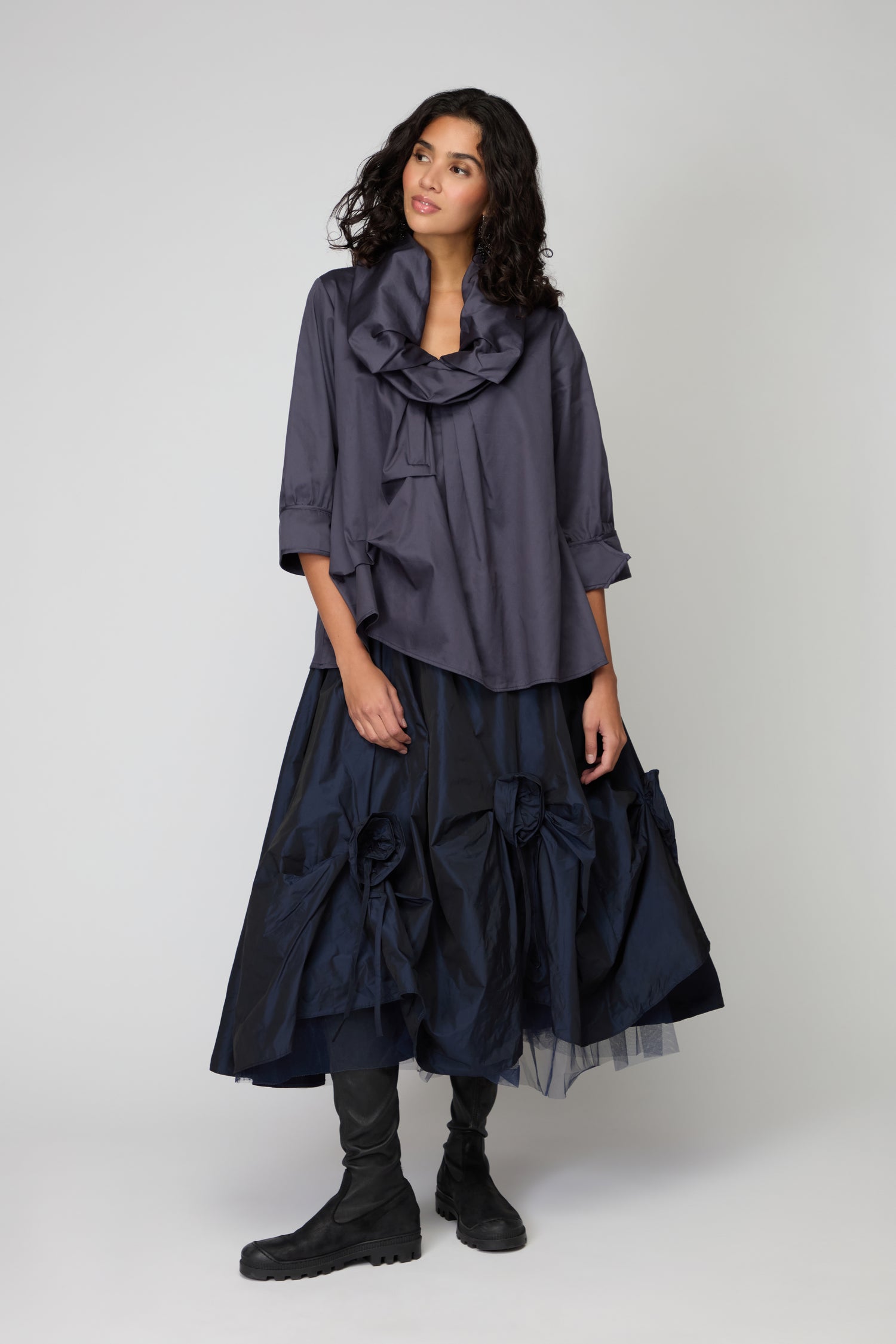 A woman stands against a plain background wearing the Frill Neck Easy Top in dark blue, featuring a ruffled collar and three-quarter sleeves, paired with a navy voluminous skirt with large bows and black boots.
