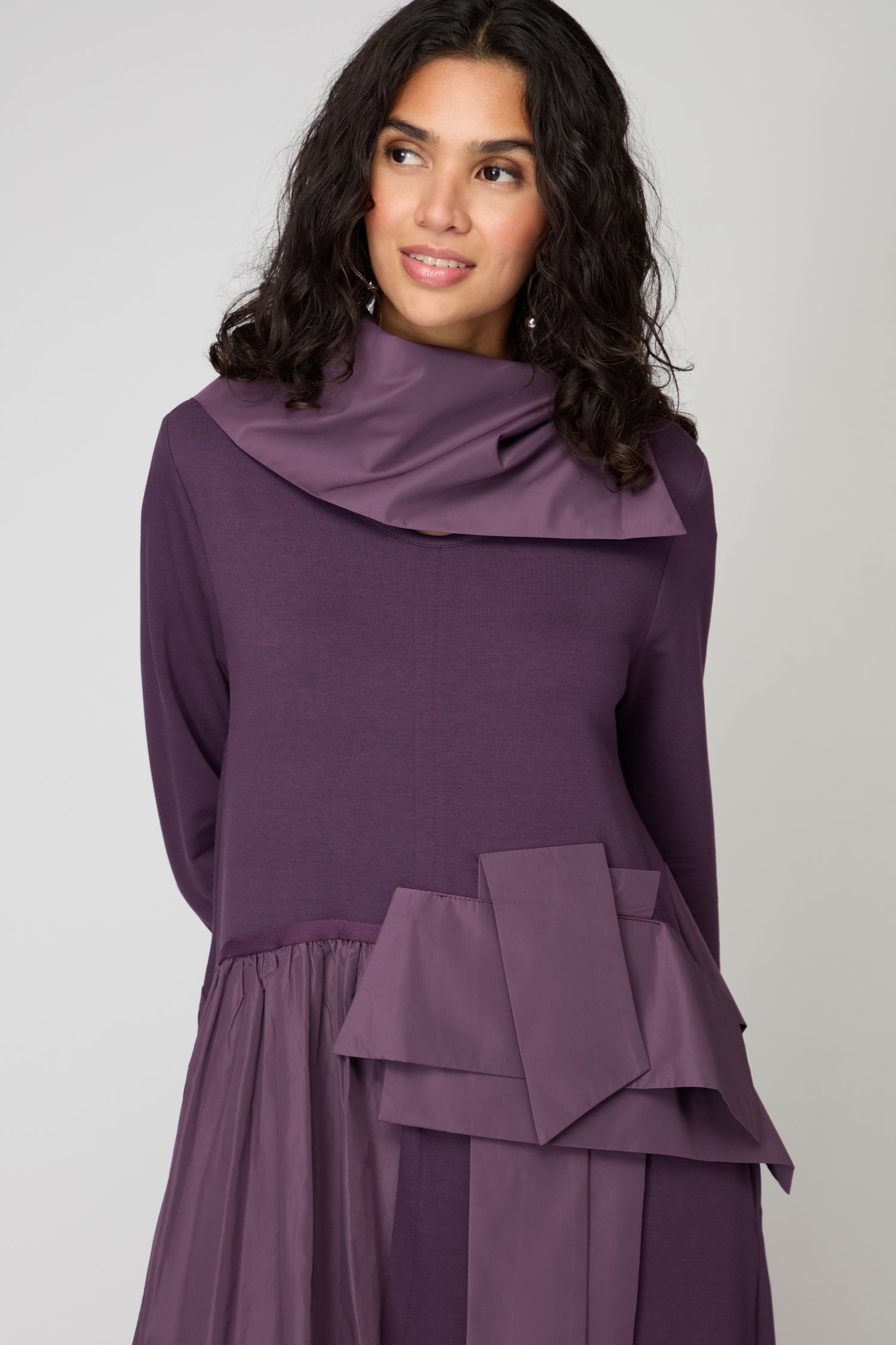 A woman with medium-length curly hair wears the Jersey Taffetta Waist Detail Dress in purple, featuring asymmetrical fabric accents, a high neckline, and a relaxed fit, standing against a plain background.