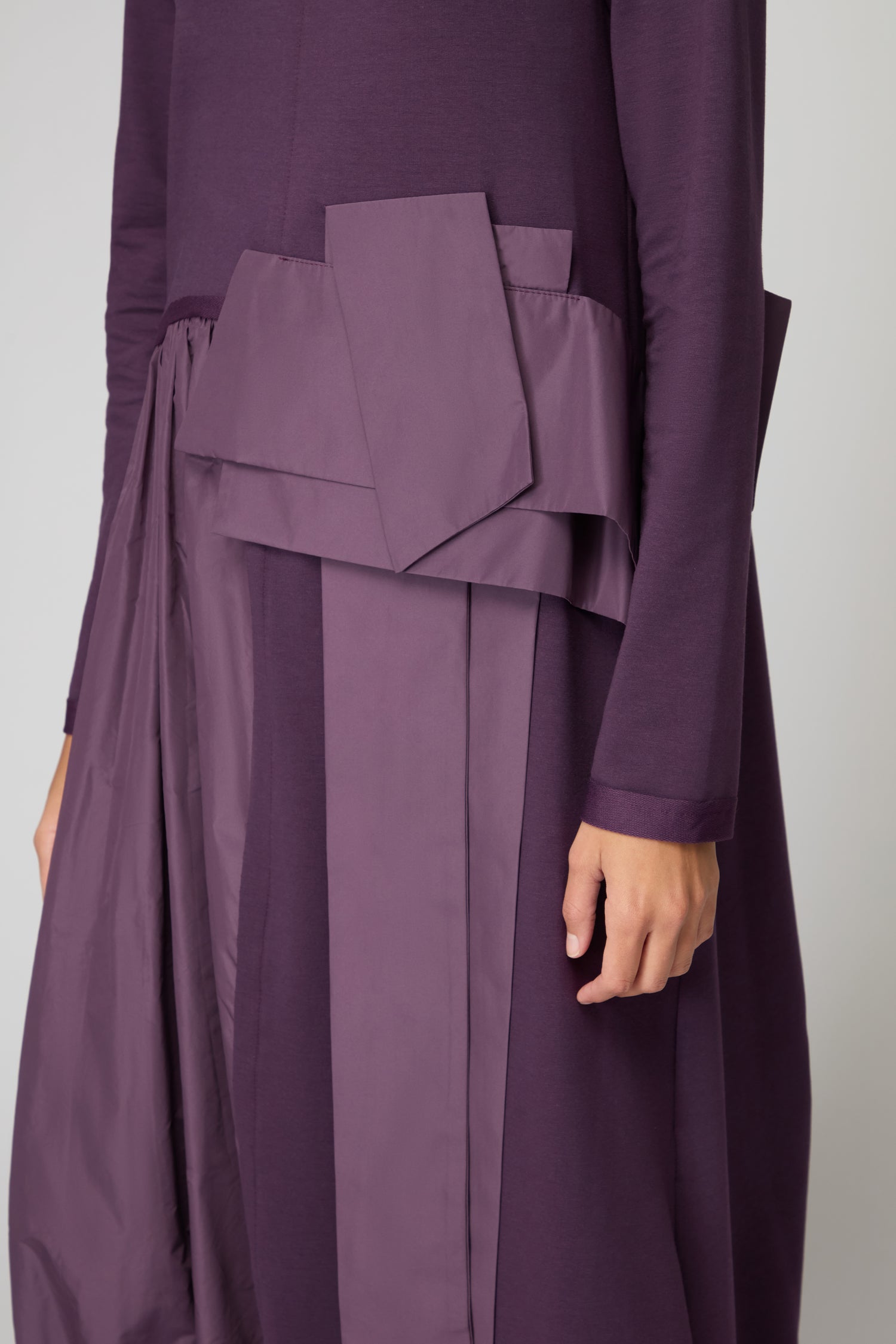 A person wears the Jersey Taffeta Waist Detail Dress, a long-sleeved, dark purple dress featuring geometric, layered fabric accents and waist panels. Only the torso and part of one arm are visible.