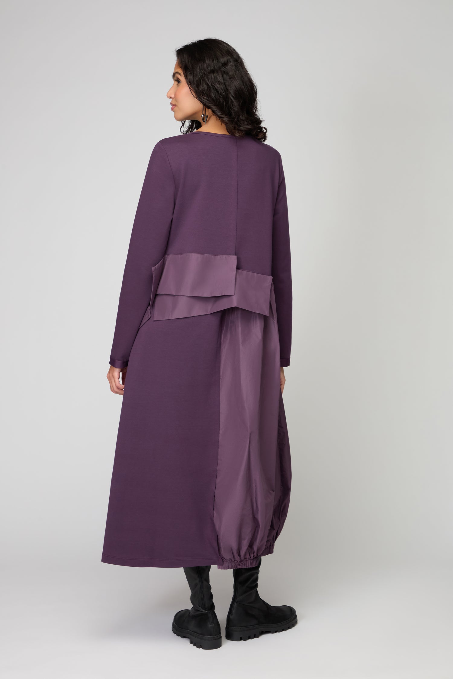 A woman stands facing away in a purple, long-sleeved Jersey Taffetta Waist Detail Dress with an ankle-length relaxed fit, satin panel, and back pleat detail, paired with black boots against a plain background.
