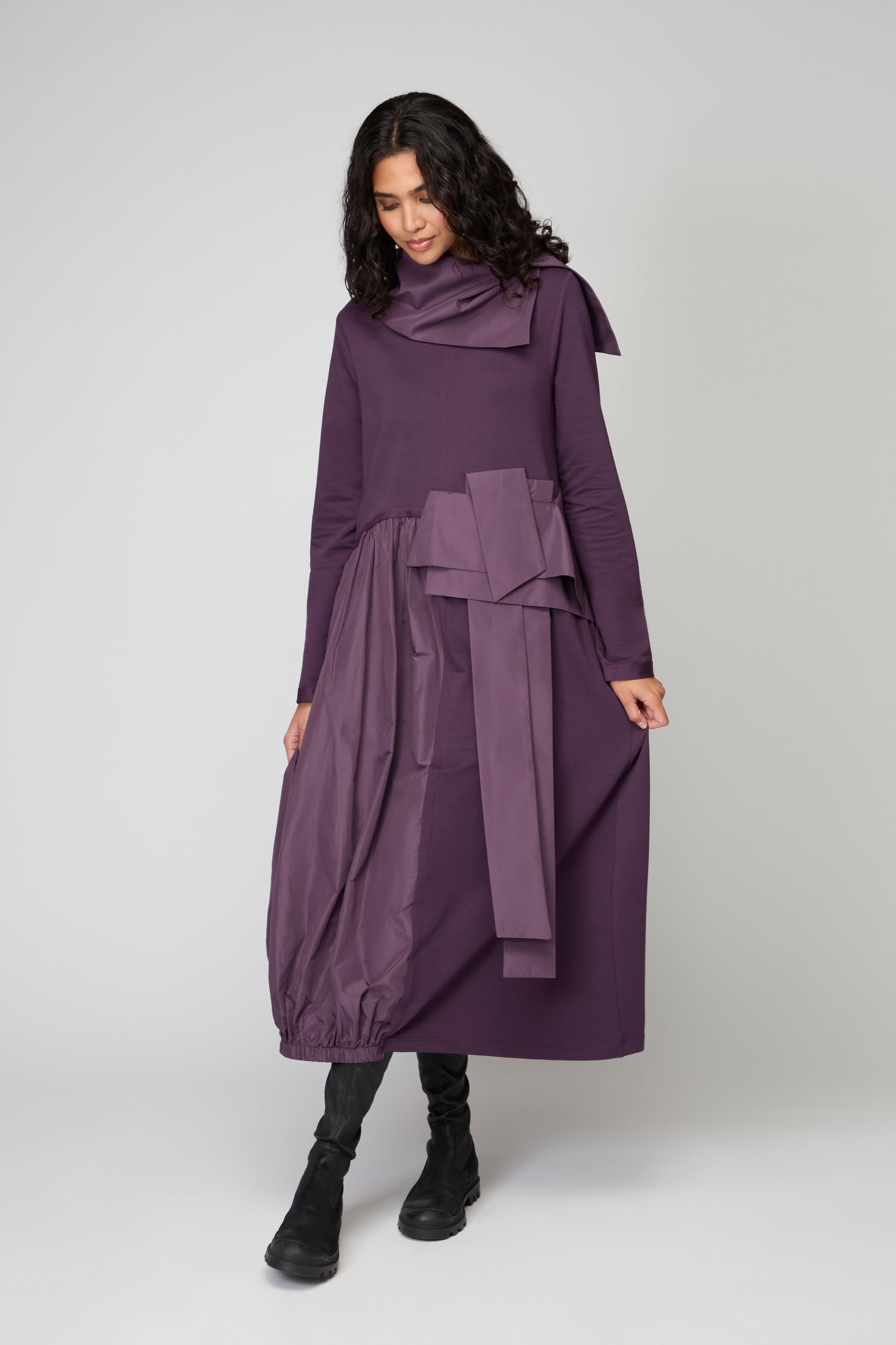 A woman in a purple Jersey Taffetta Waist Detail Dress featuring long sleeves, a relaxed fit, and an asymmetric bow stands against a plain background, looking down and holding the skirt. She pairs the dress with black boots.
