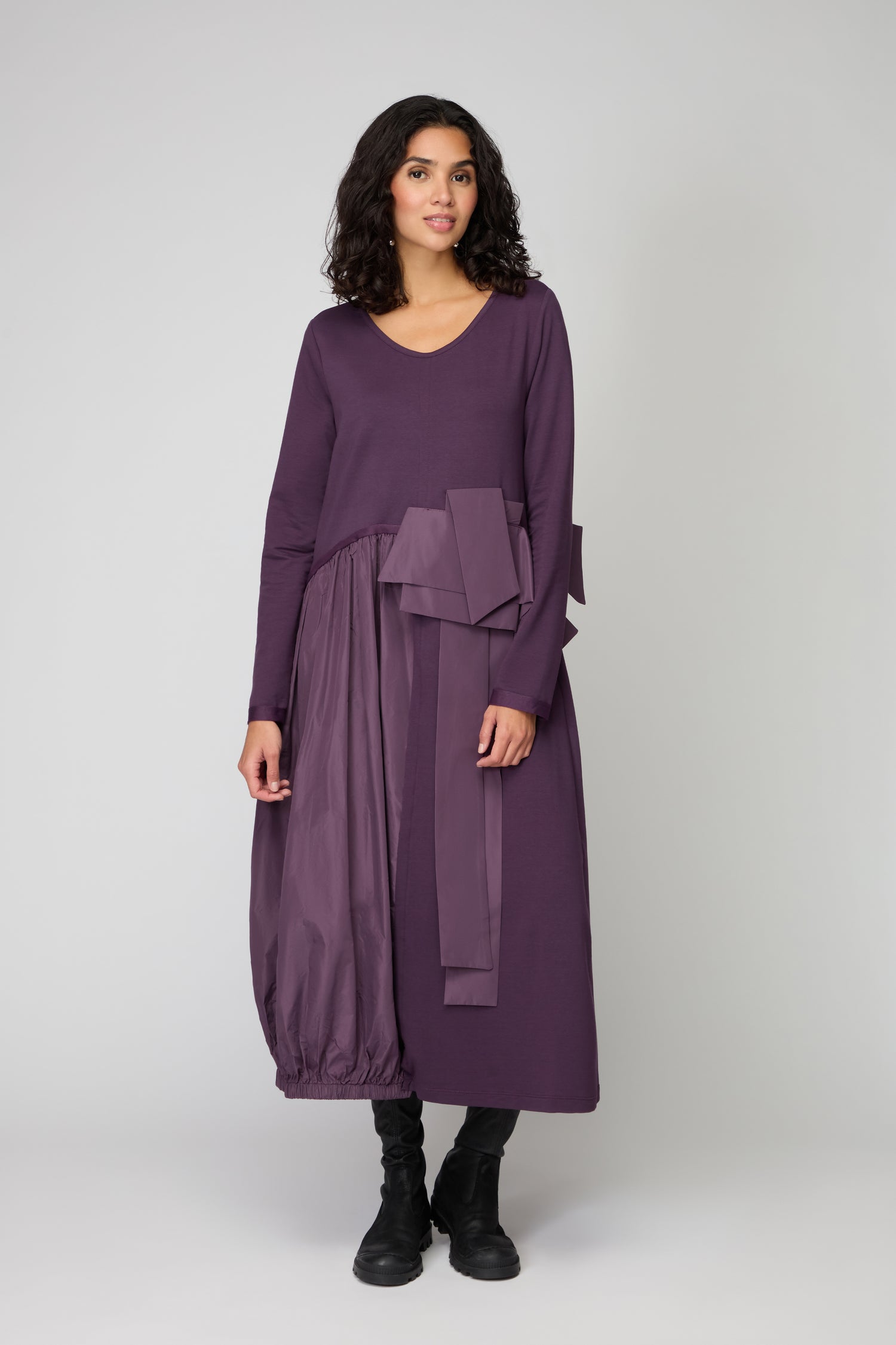 A woman poses against a plain background wearing the Jersey Taffeta Waist Detail Dress featuring a relaxed fit, long sleeves, an asymmetrical skirt, bow detail, and paired with black boots.