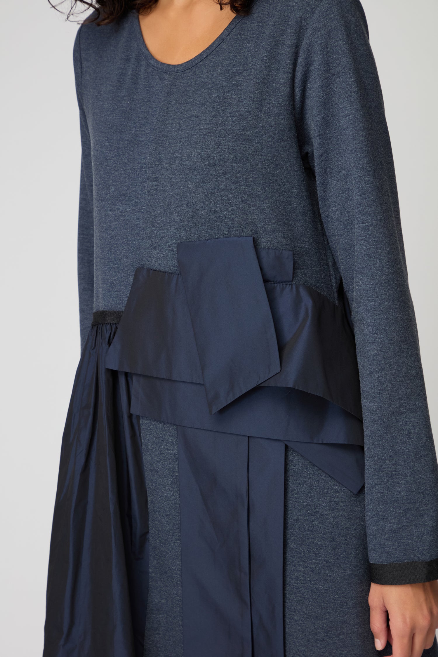 A person wears the Jersey Taffetta Waist Detail Dress, featuring long sleeves, blue taffeta fabric with layered asymmetrical details, a relaxed silhouette, and a bow accent at the waist.