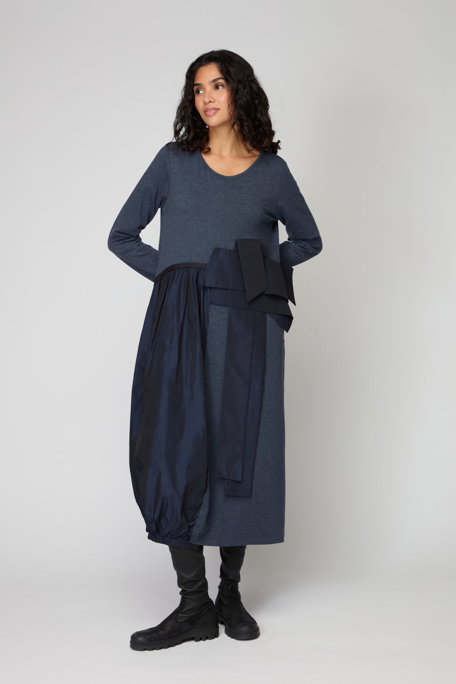 A woman stands against a plain background wearing the Jersey Taffetta Waist Detail Dress with a relaxed fit, asymmetrical navy accents, and black boots.