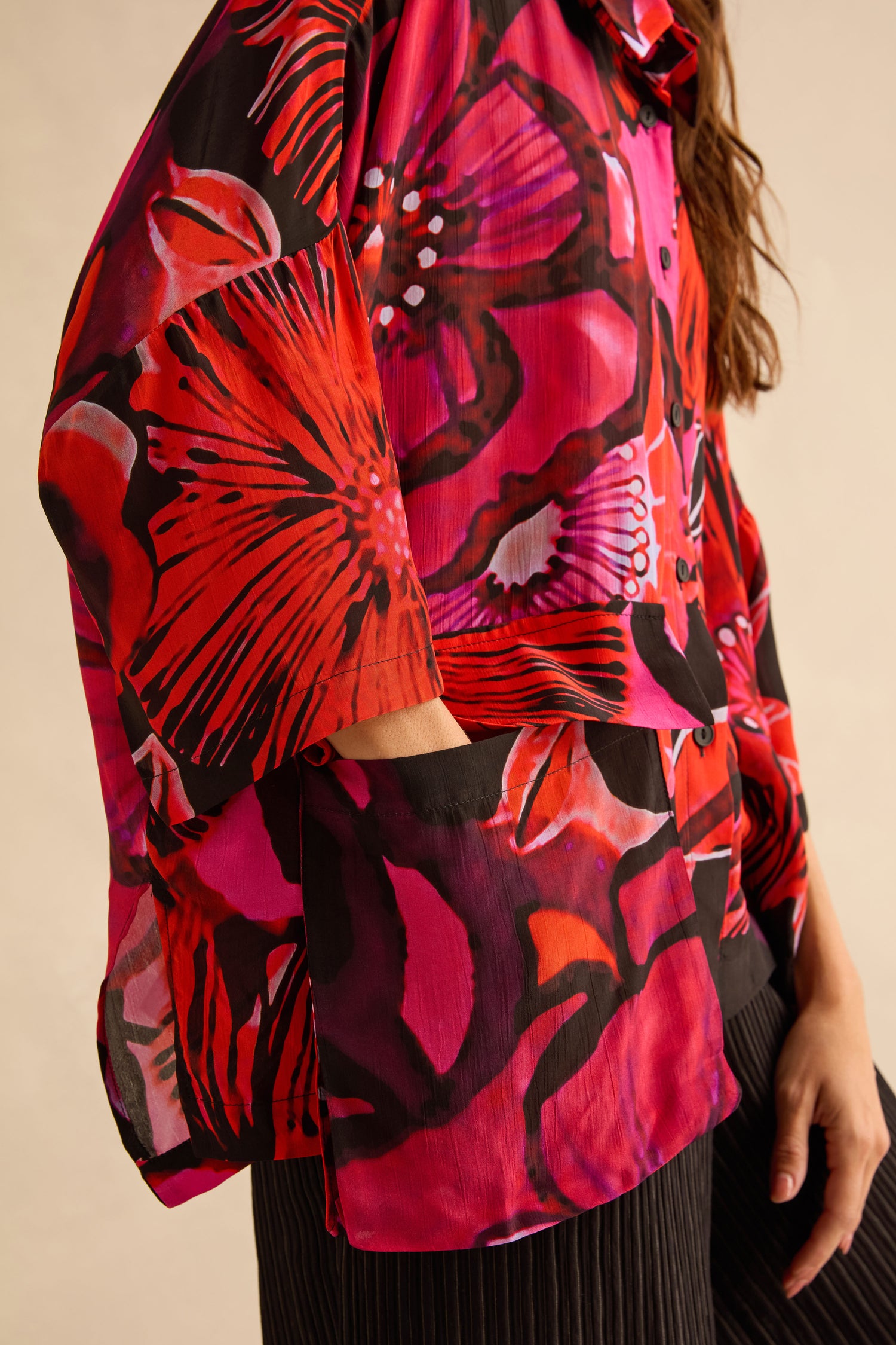 A person wearing a Pocket Floral Shirt made of viscose, featuring a short-sleeved, button-up design with a bold red, pink, and black floral print and a front patch pocket.
