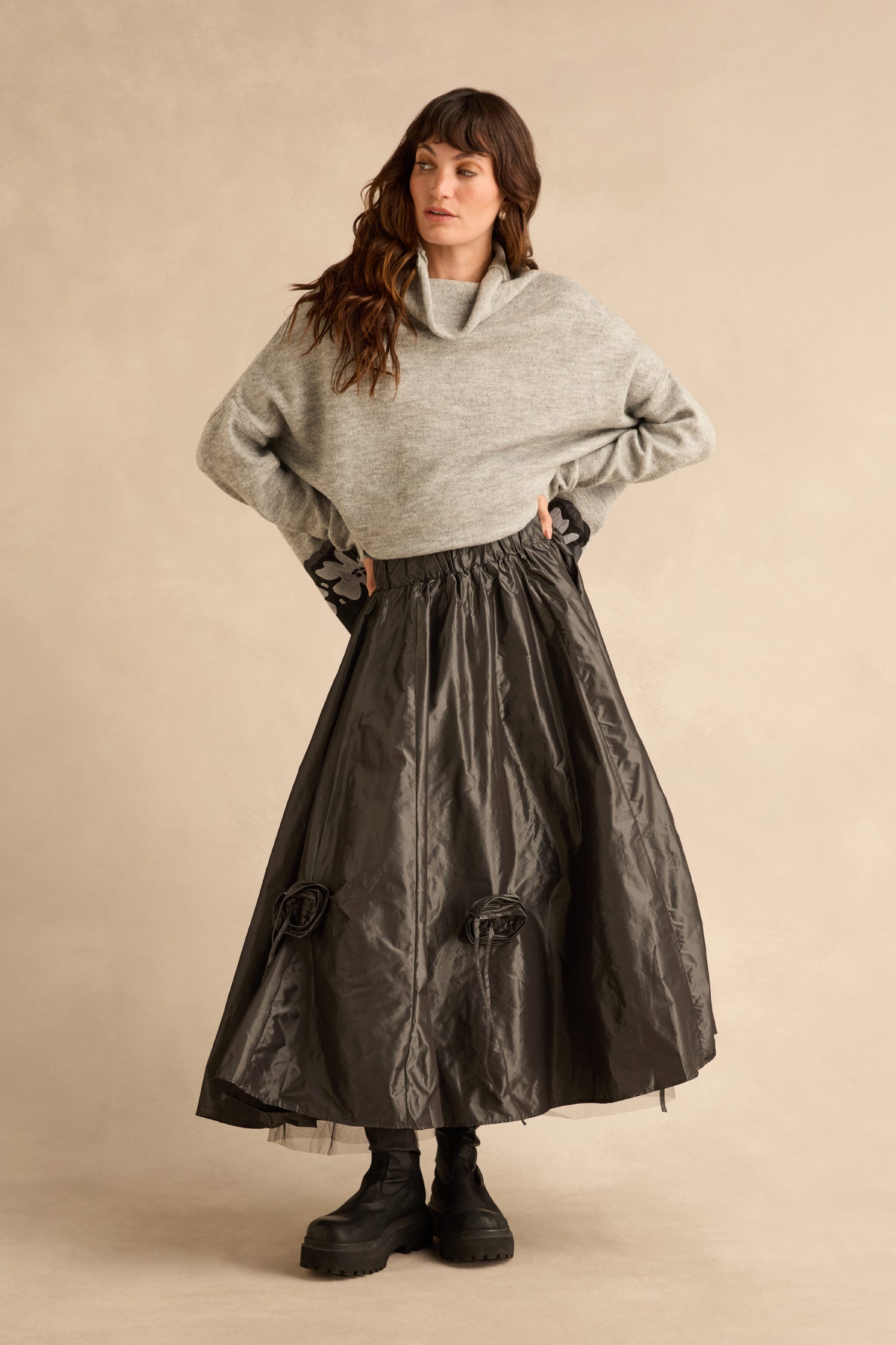 A person wearing a metallic grey cowl-neck sweater, a Flower Detail Taffeta Skirt with fabric flower accents, and black platform boots.