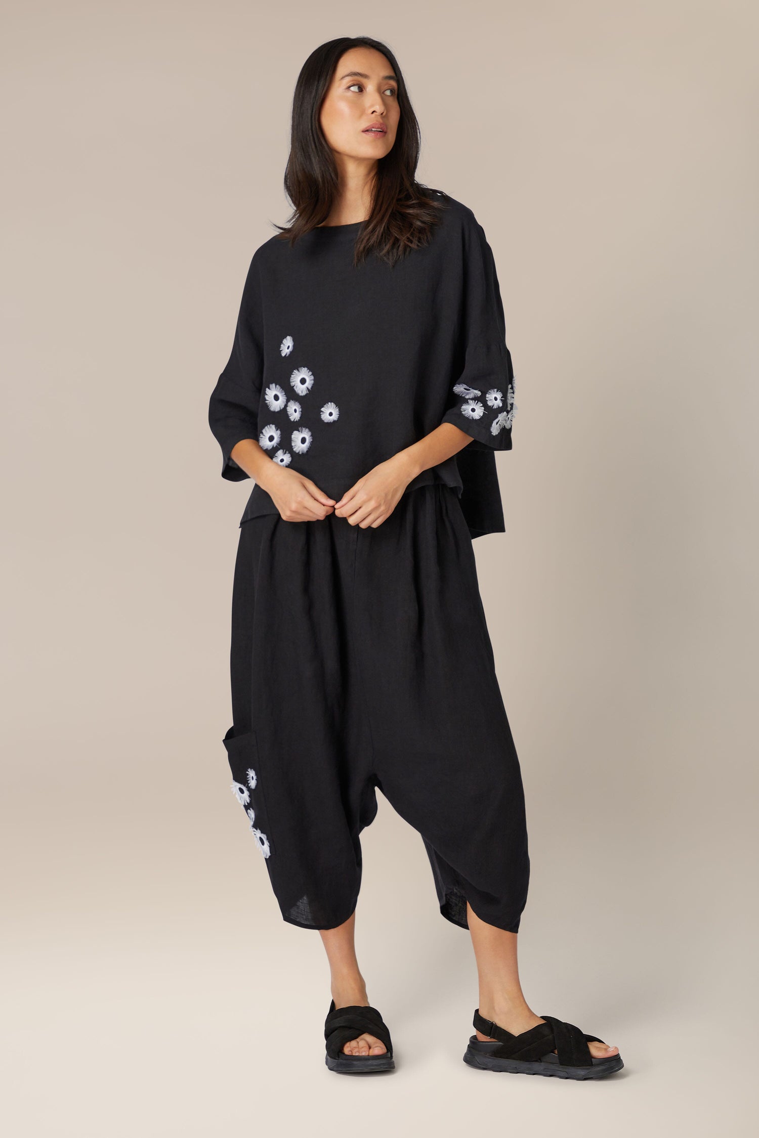 Woman in black outfit with Magnolia Linen Top, featuring floral embroidery and a high boat neck, posing on a neutral background.