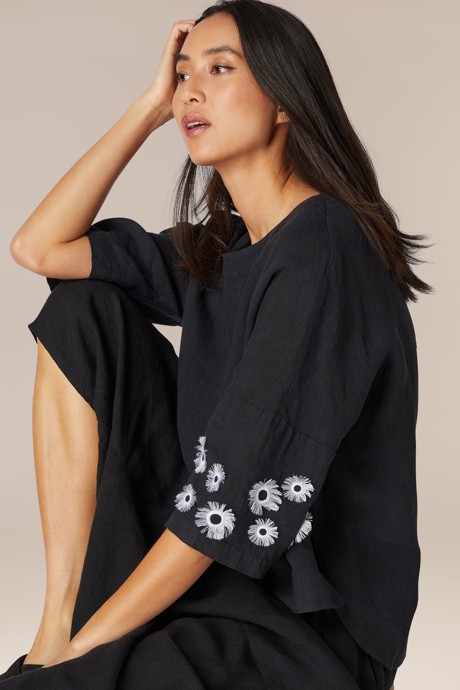 Woman in a Sunflower Linen Top with high boat neck and avant-garde design, featuring floral embroidery on the sleeves, posing with her hand on her head.
