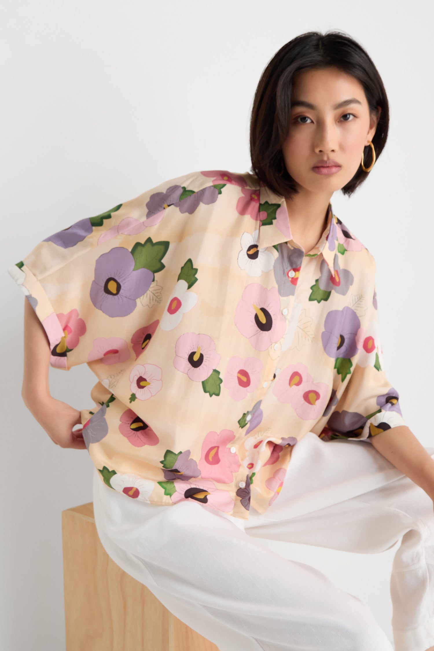 A person with short dark hair wears the Rose Eclipse Silk Boxy Shirt and white pants, sitting on a wooden block against a plain white background.