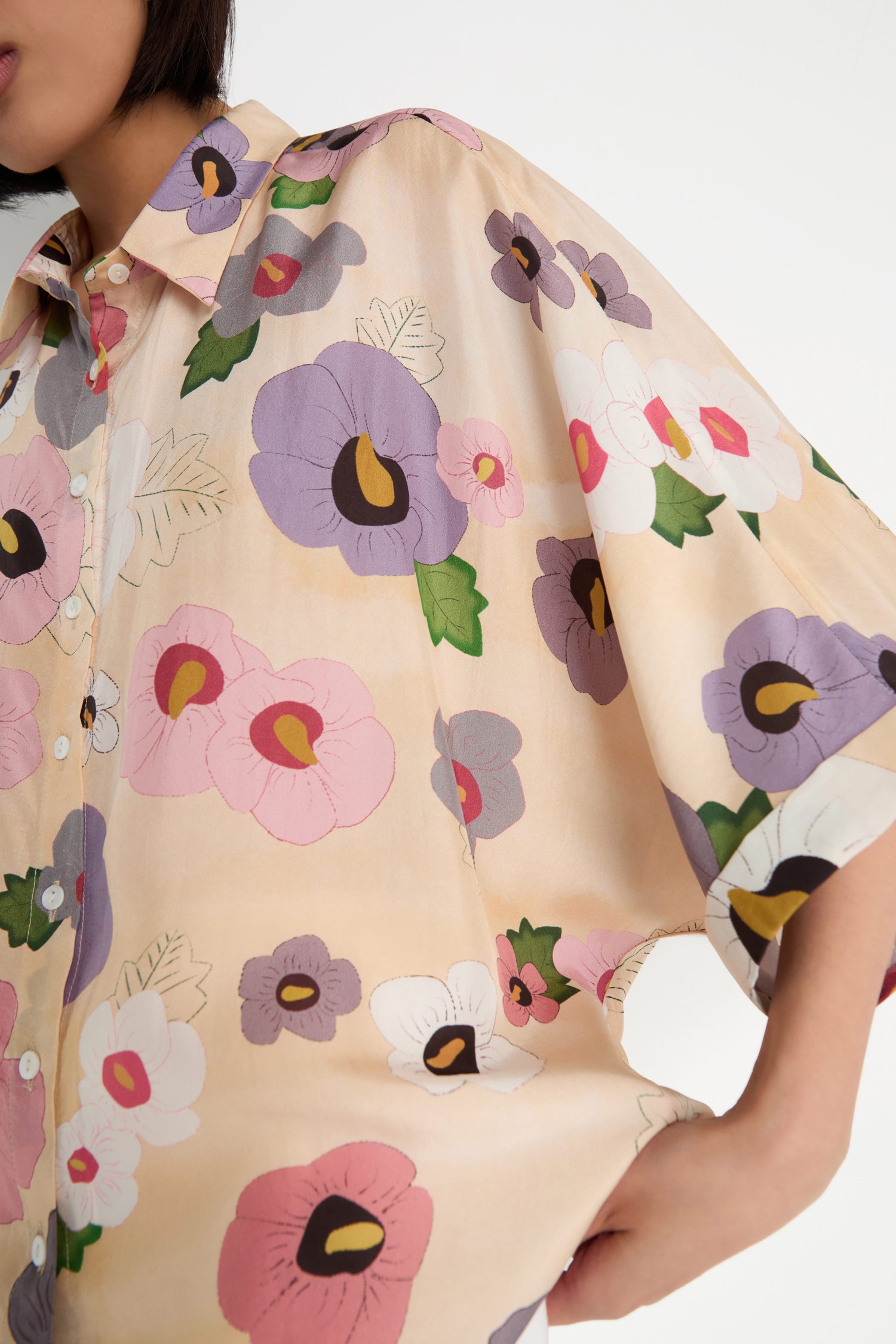 A person wears the Rose Eclipse Silk Boxy Shirt—a loose, light-colored top with large, colorful floral graphics in pink, purple, green, and white. Only their upper body and part of an arm are visible.